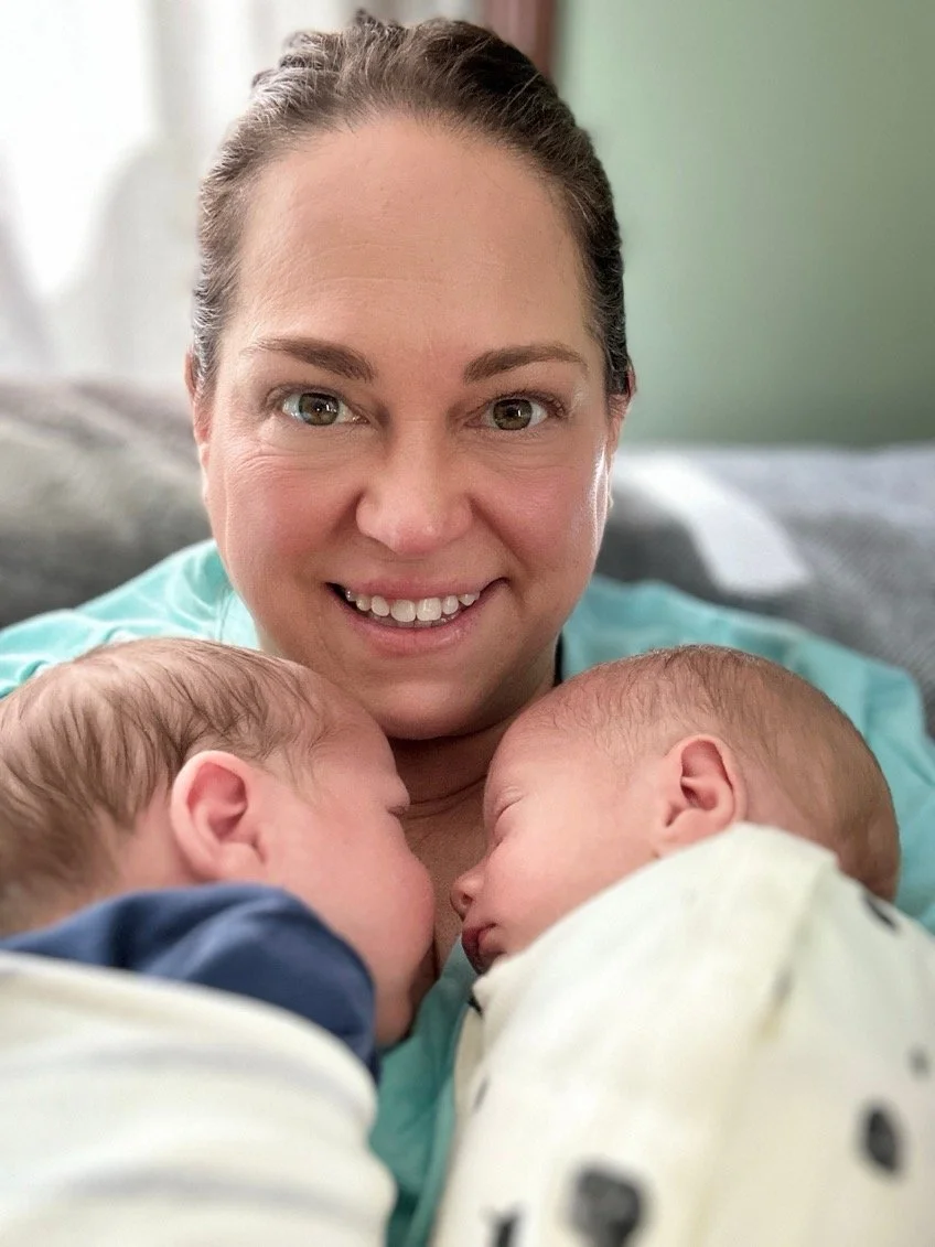A woman smiling while holding two sleeping babies close to her chest, with their faces touching.