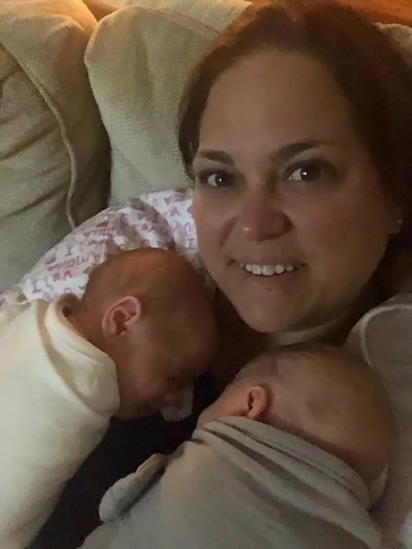 Two babies lying on a bed with animal-themed bedsheets. One baby is waving while the other looks at the camera.