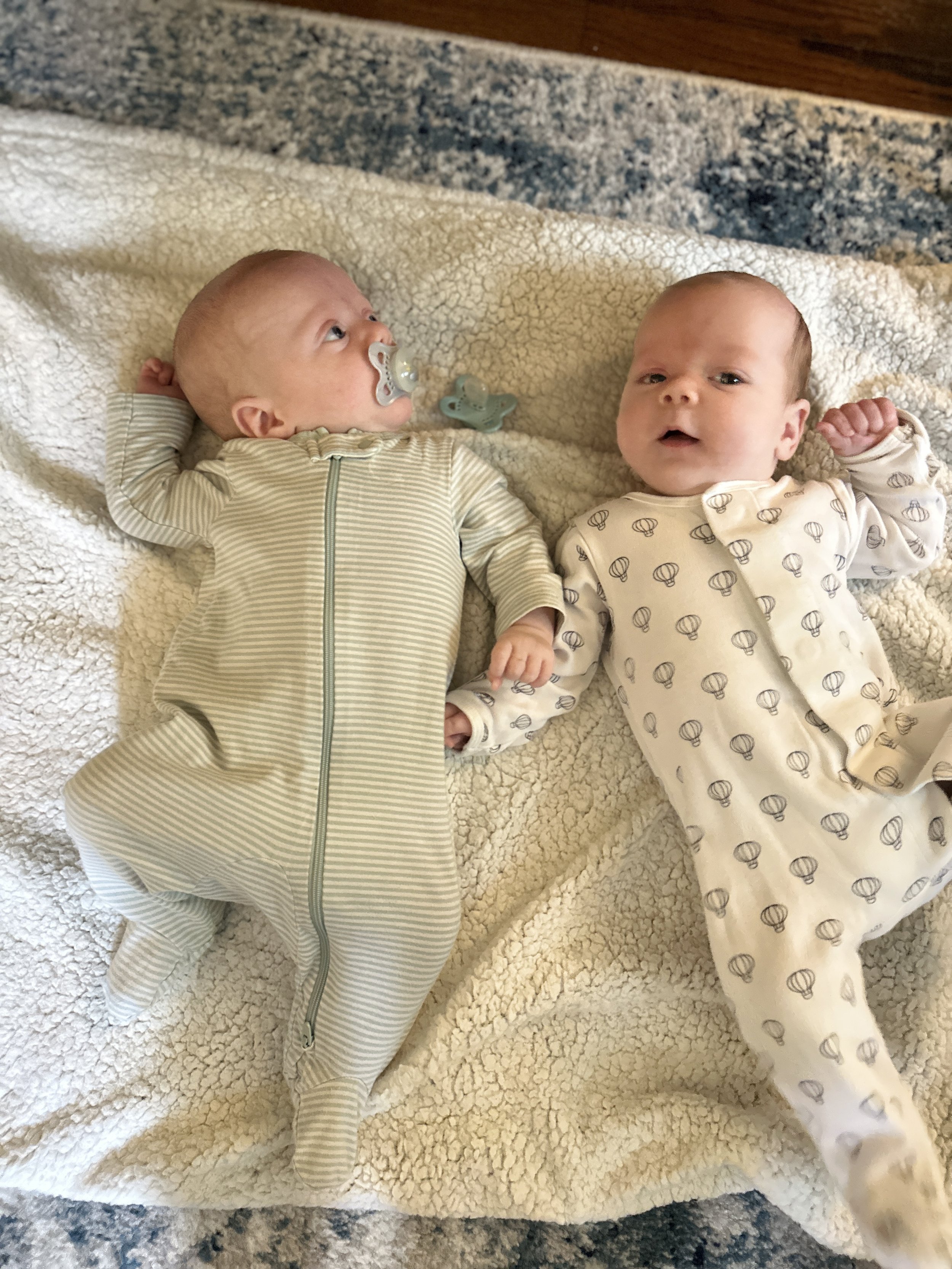 Two babies lying on a blanket, one with a pacifier and the other looking at the camera, both wearing cream-colored outfits with different patterns.