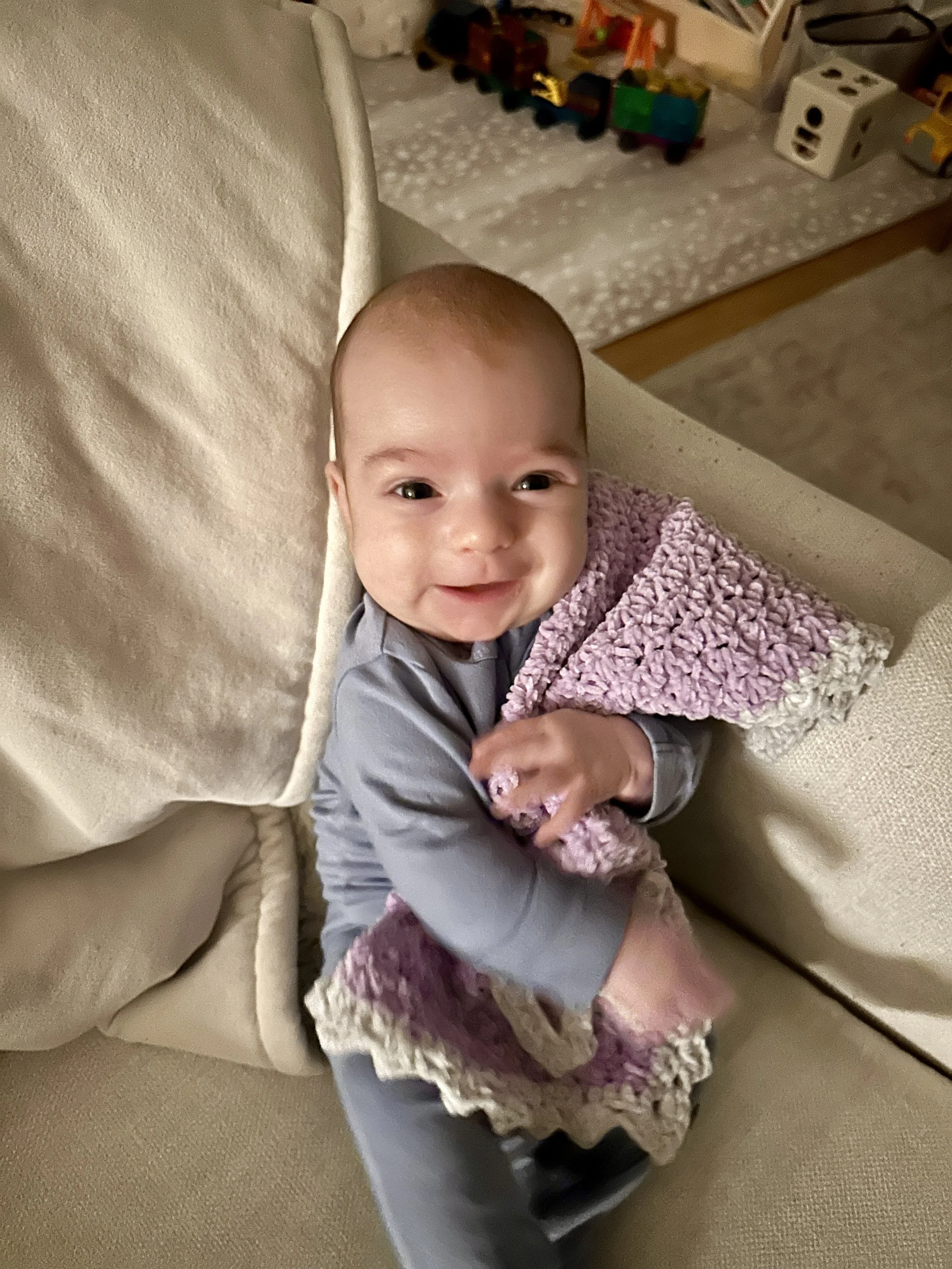 Smiling baby sitting on a beige couch holding a pink and white crocheted blanket.