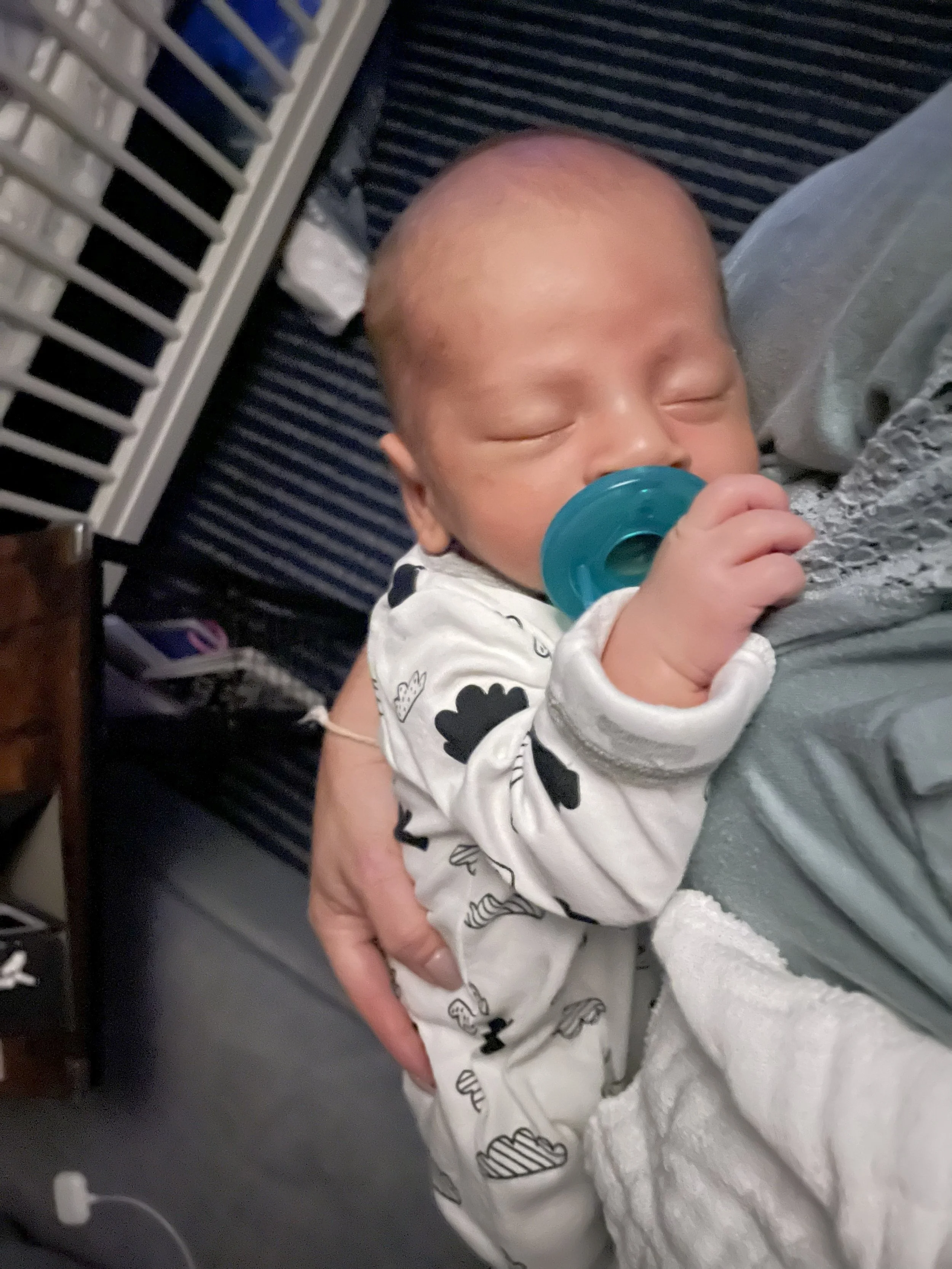 A sleeping baby with a pacifier, lying on an adult's chest, wearing a white onesie with black cartoon faces and clouds, with a hospital or clinical setting background.