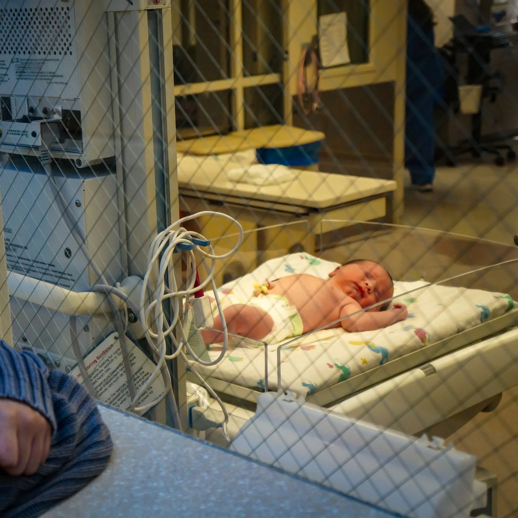 Newborn baby lying in a hospital incubator, connected to medical equipment through wires, in a neonatal intensive care unit.