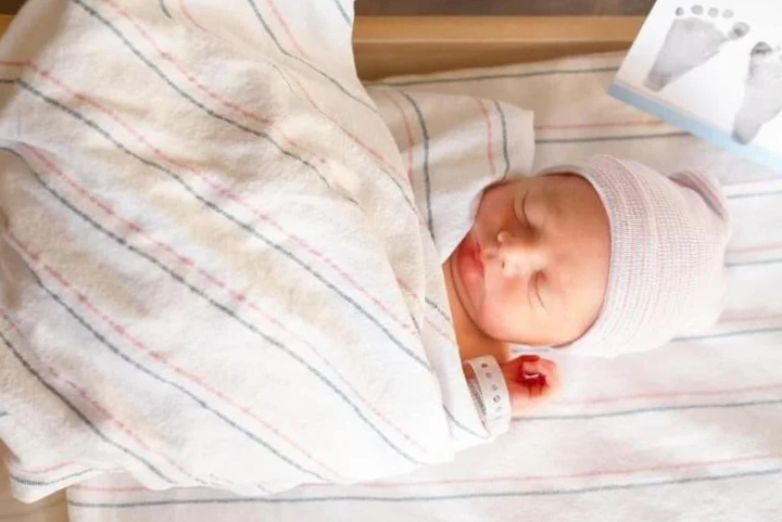 Newborn baby wrapped in hospital blanket, wearing a striped hospital hat, lying in a hospital bed with a photo of footprints in the top right corner.