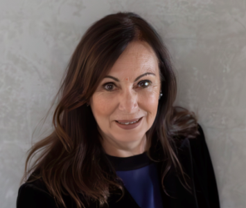 A woman with shoulder-length brown hair, smiling, wearing a black top and earrings, standing against a gray textured background.