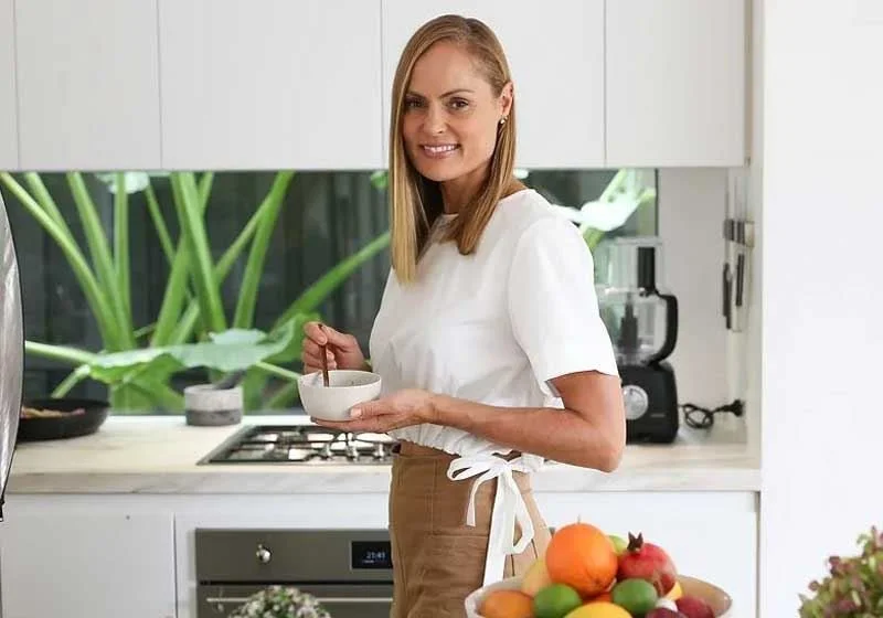 Woman smiling in a kitchen holding a bowl with a spoon, wearing a white top and apron, with a bowl of assorted fruits on the counter.
