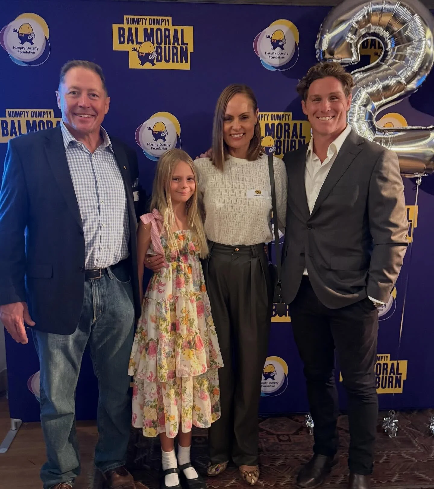 A group of four people, three adults and one young girl, posing together at an event with a purple backdrop featuring logos and banners for 'Humpty Dumpty Foundation' and 'Balmoral Burn.' There is a large silver balloon shaped like the number 2 beside them.