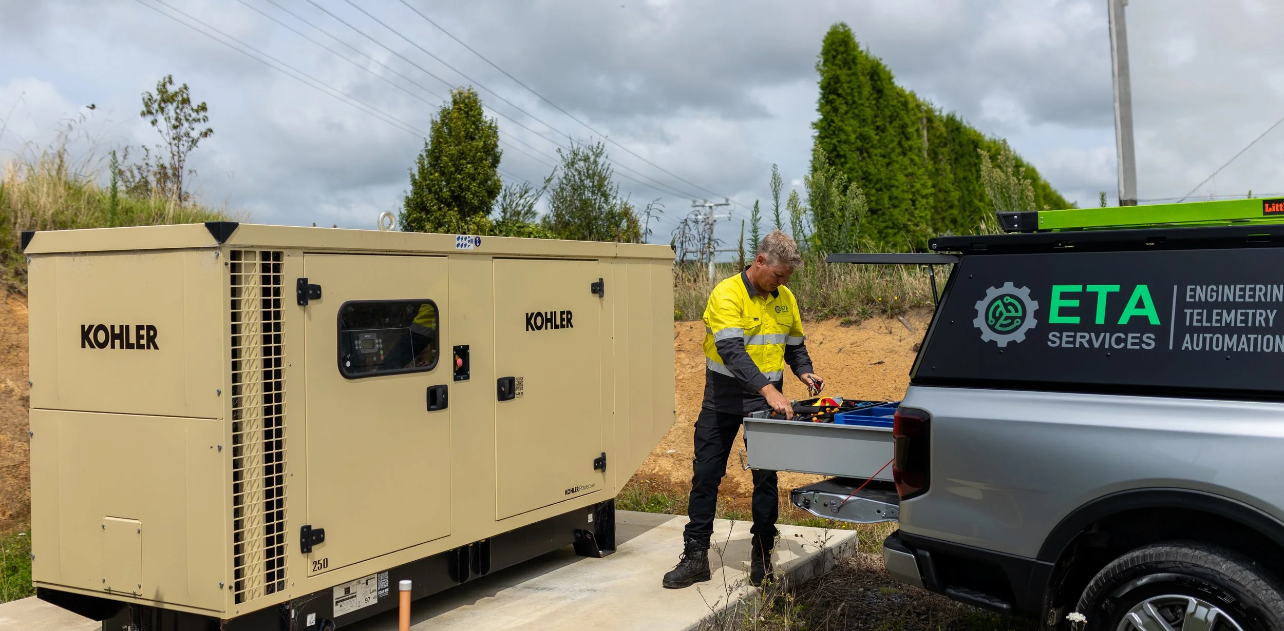 A technician in a yellow and black jacket working with equipment from a service truck parked outdoors, near a large Kohler generator on a concrete slab, with trees and a cloudy sky in the background.