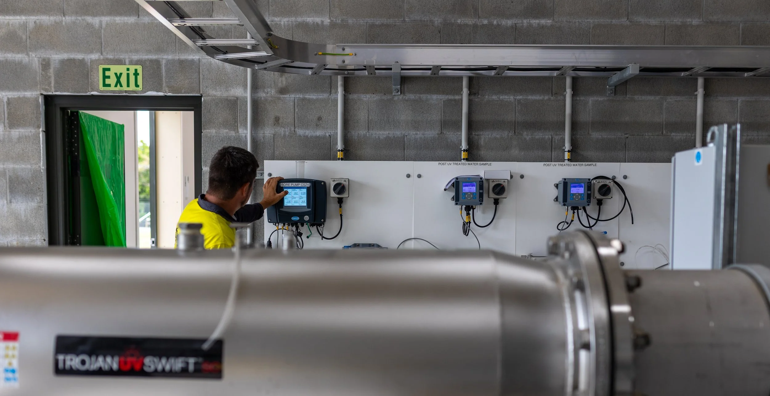 A technician adjusts a control panel on a water treatment system, with water sample testing devices mounted on a white panel labeled for post UV treated water sample, inside a utility room with concrete walls and an exit sign.
