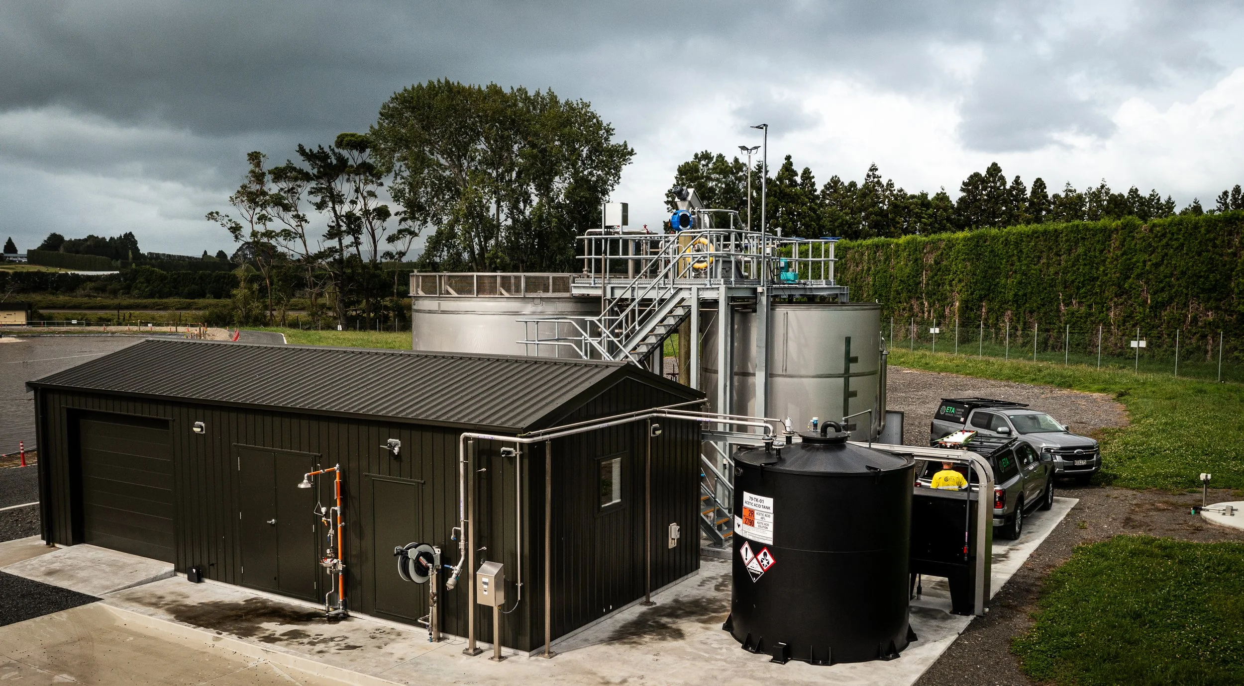 An industrial wastewater treatment facility with black and gray tanks, metal walkways, and vehicles parked beside the equipment, set outdoors with a hedge and trees in the background under a cloudy sky.