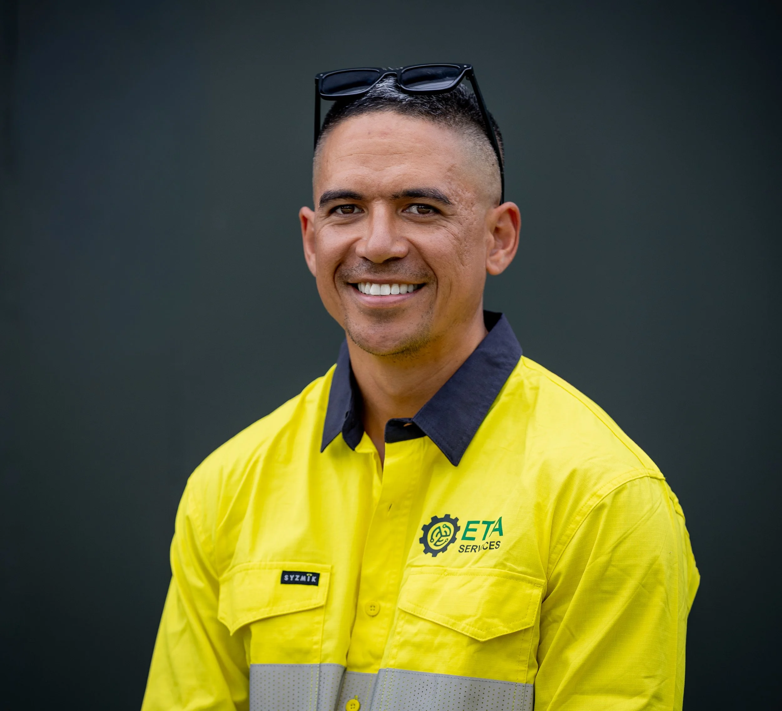 Smiling man wearing yellow uniform with ETA Services logo, sunglasses on head, against dark background.