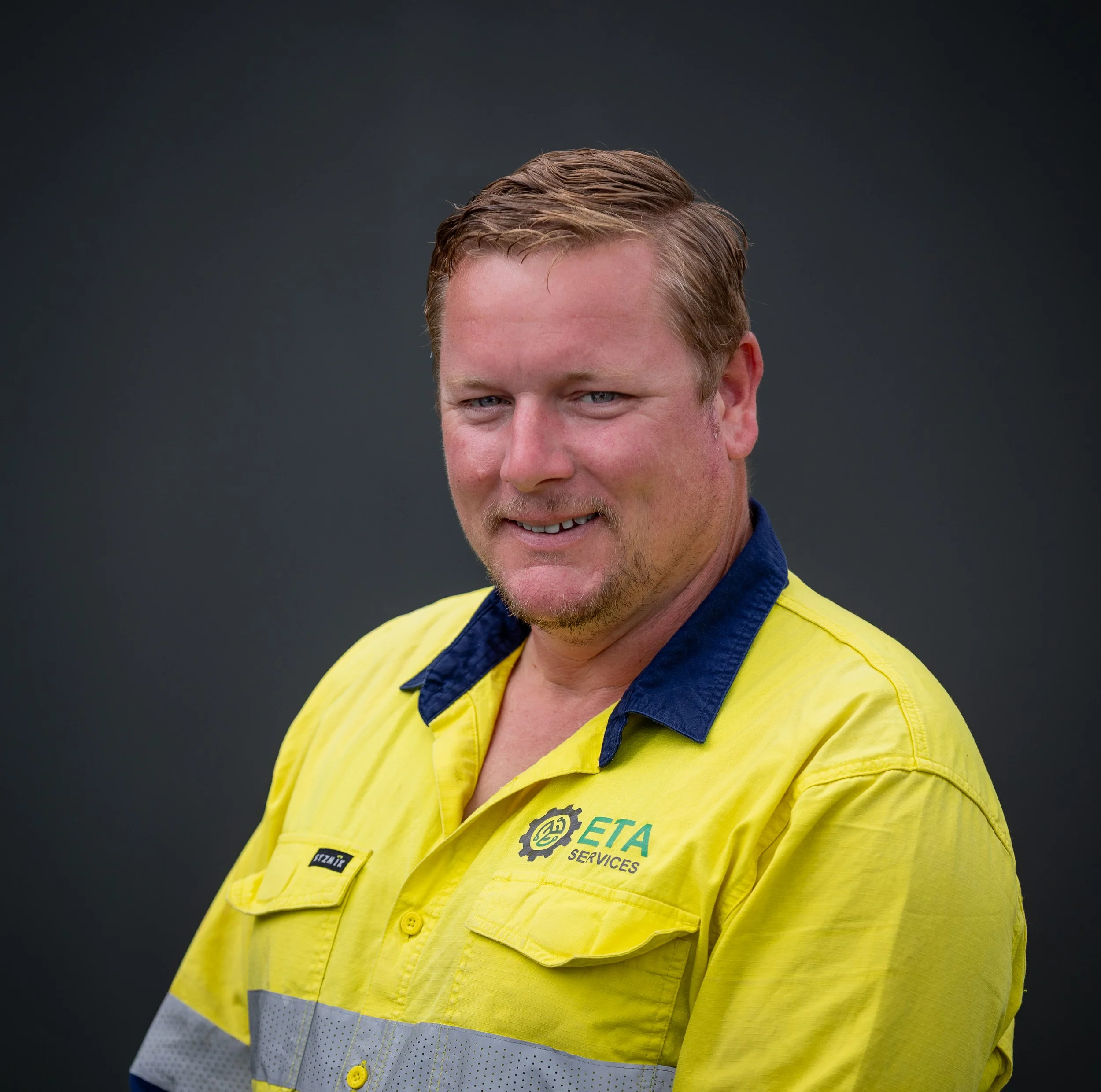 A man with blonde hair and a goatee smiles while wearing a yellow and navy work shirt with ETA Services logo against a dark background.