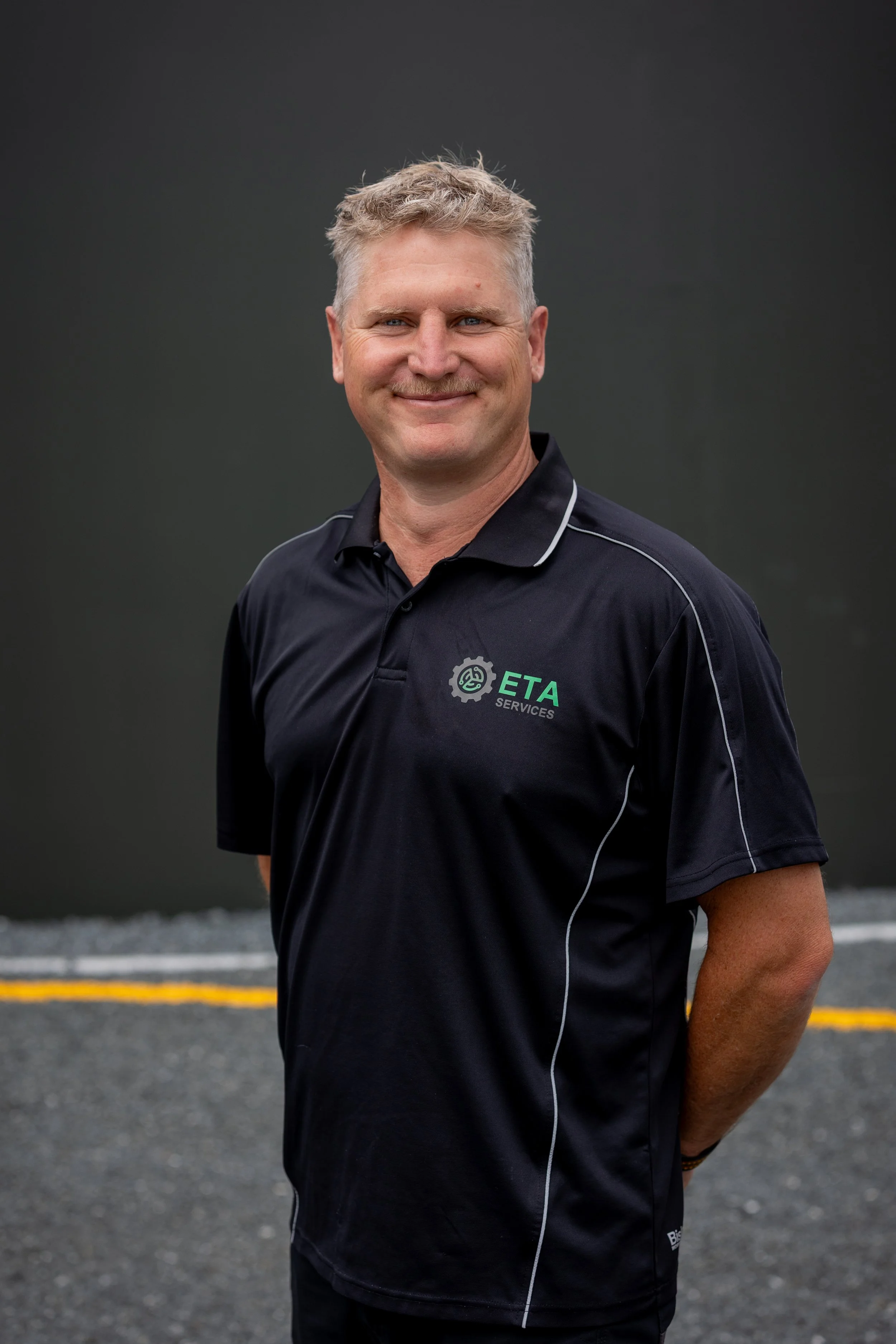 A smiling man with short, wavy blonde hair wearing a black polo shirt with a company logo that reads "ETA Services" standing outdoors on a paved surface against a dark background.