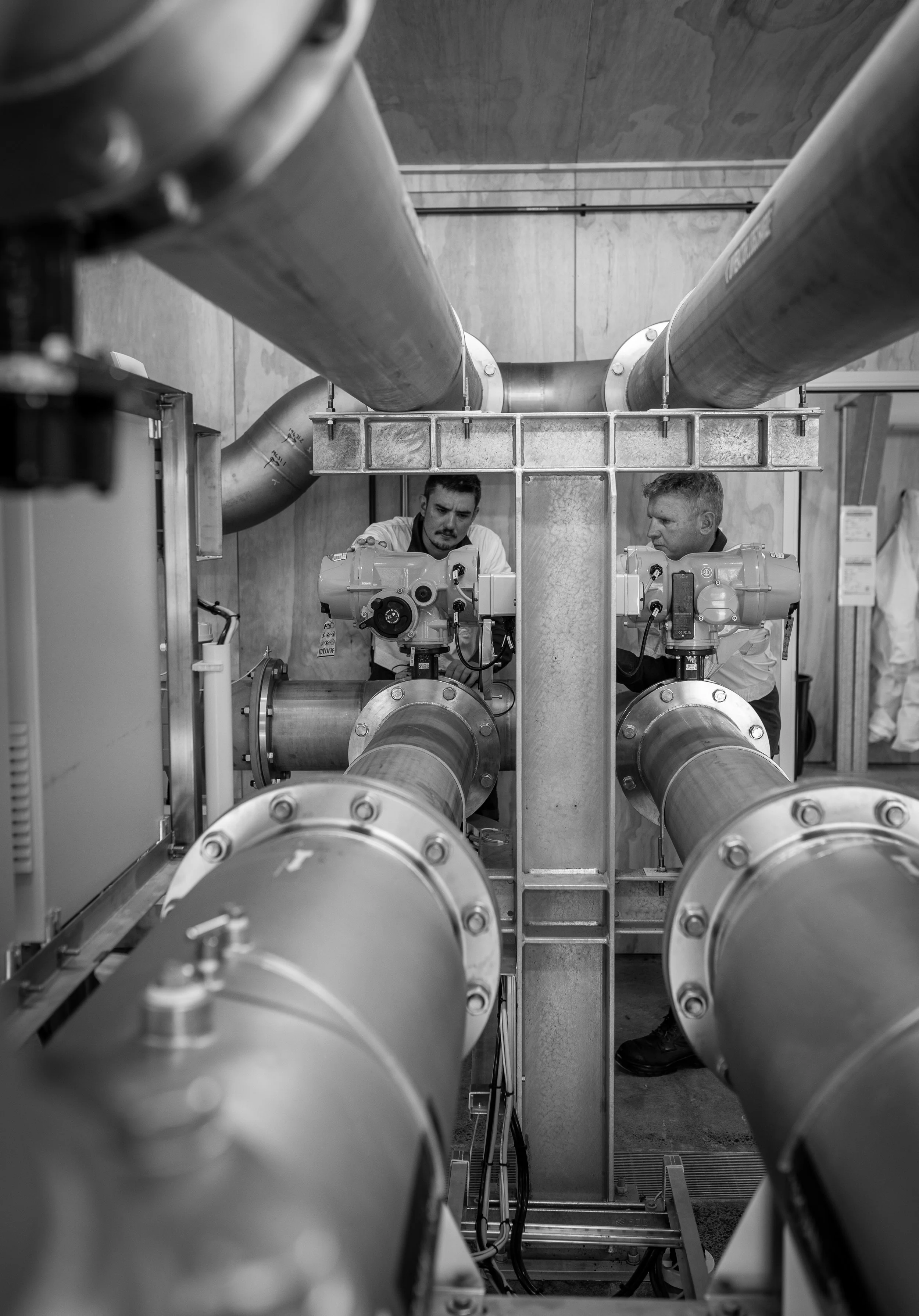 Two men operating industrial machinery with large pipes in a mechanical room.
