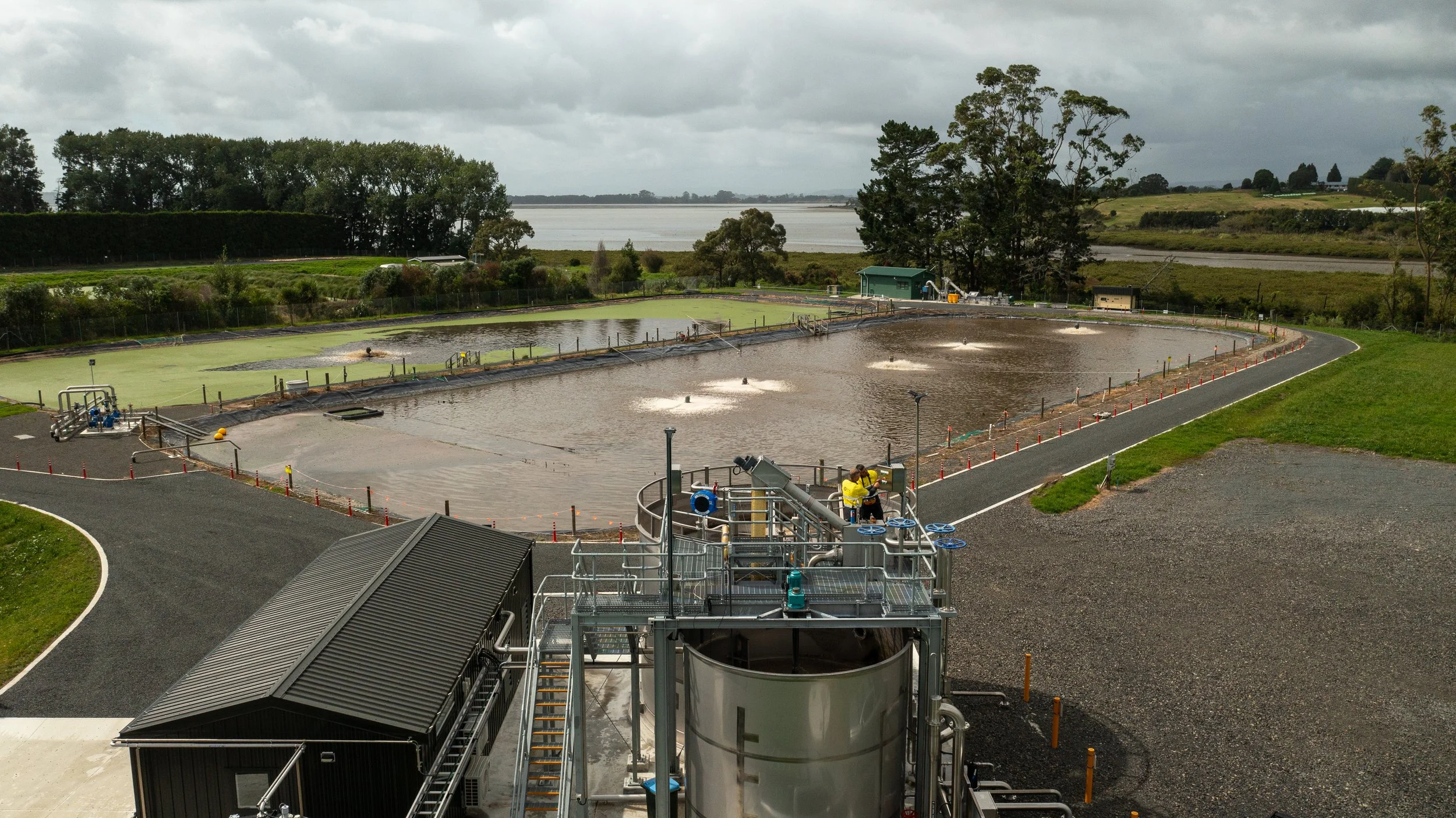 Water treatment plant with large rectangular tanks and pipes, surrounded by green fields and trees, under a cloudy sky.