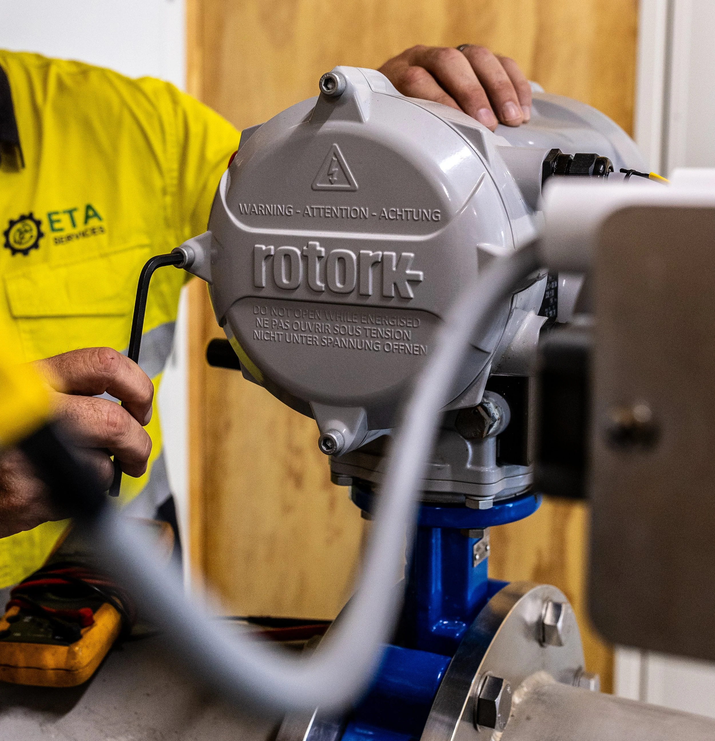 Close-up of a person handling a Rotork actuator, a gray industrial device with warning labels, connected to a blue valve, in an industrial setting.