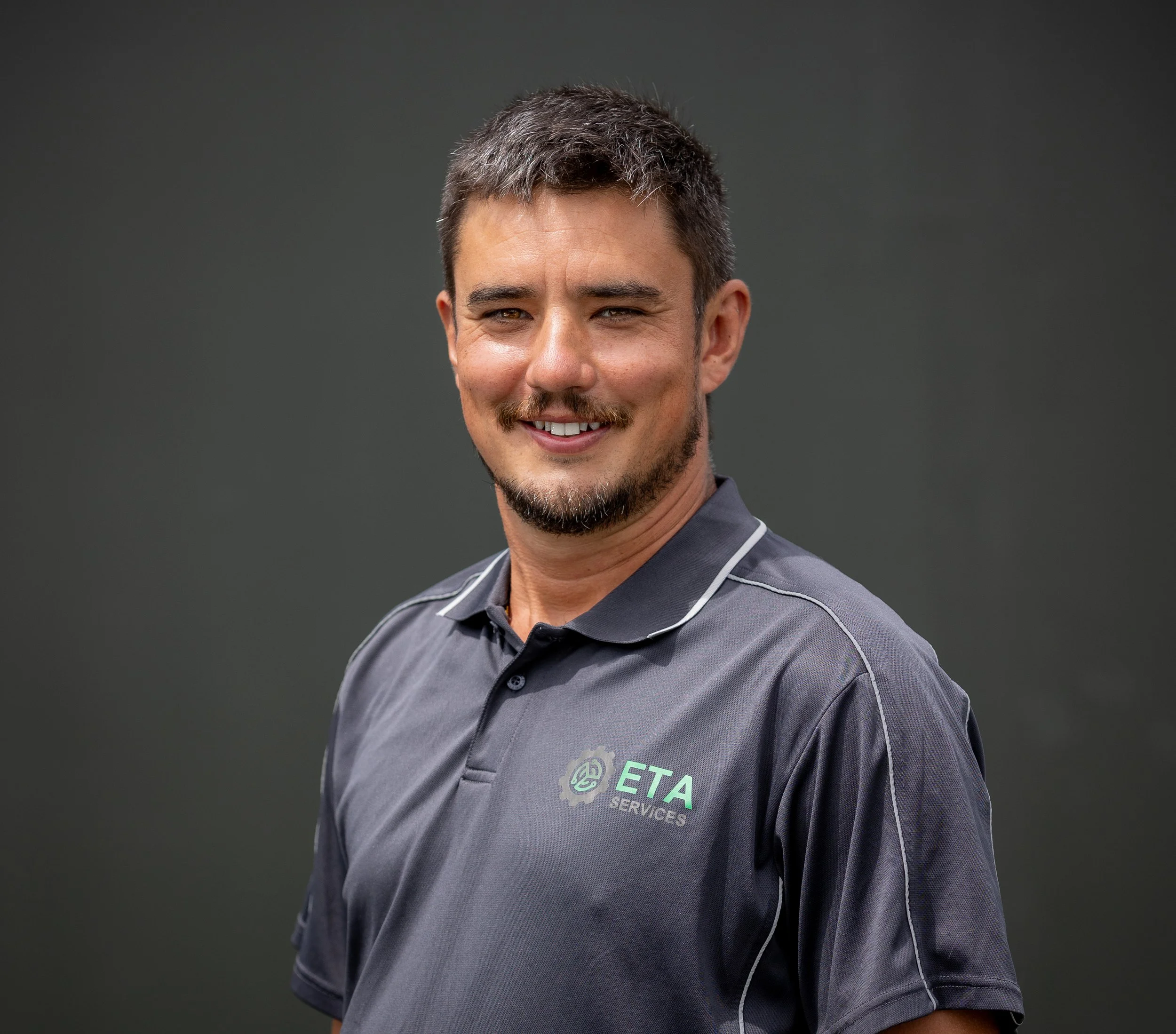A man with short dark hair and a beard, smiling, wearing a gray polo shirt with the ETA SERVICES logo, standing against a dark gray background.