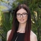 Young woman with long dark hair and glasses, smiling outdoors with greenery in the background.