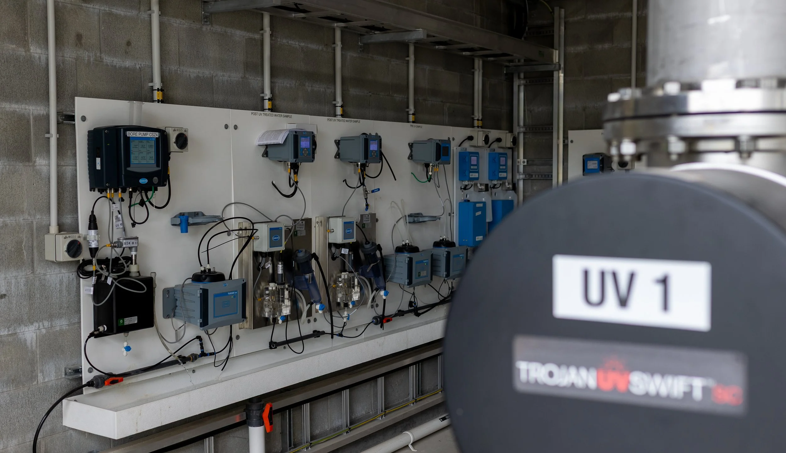 Industrial UV equipment with a sign labeled 'UV 1' in the foreground, including various electronic devices and control panels mounted on a wall in a utility room.