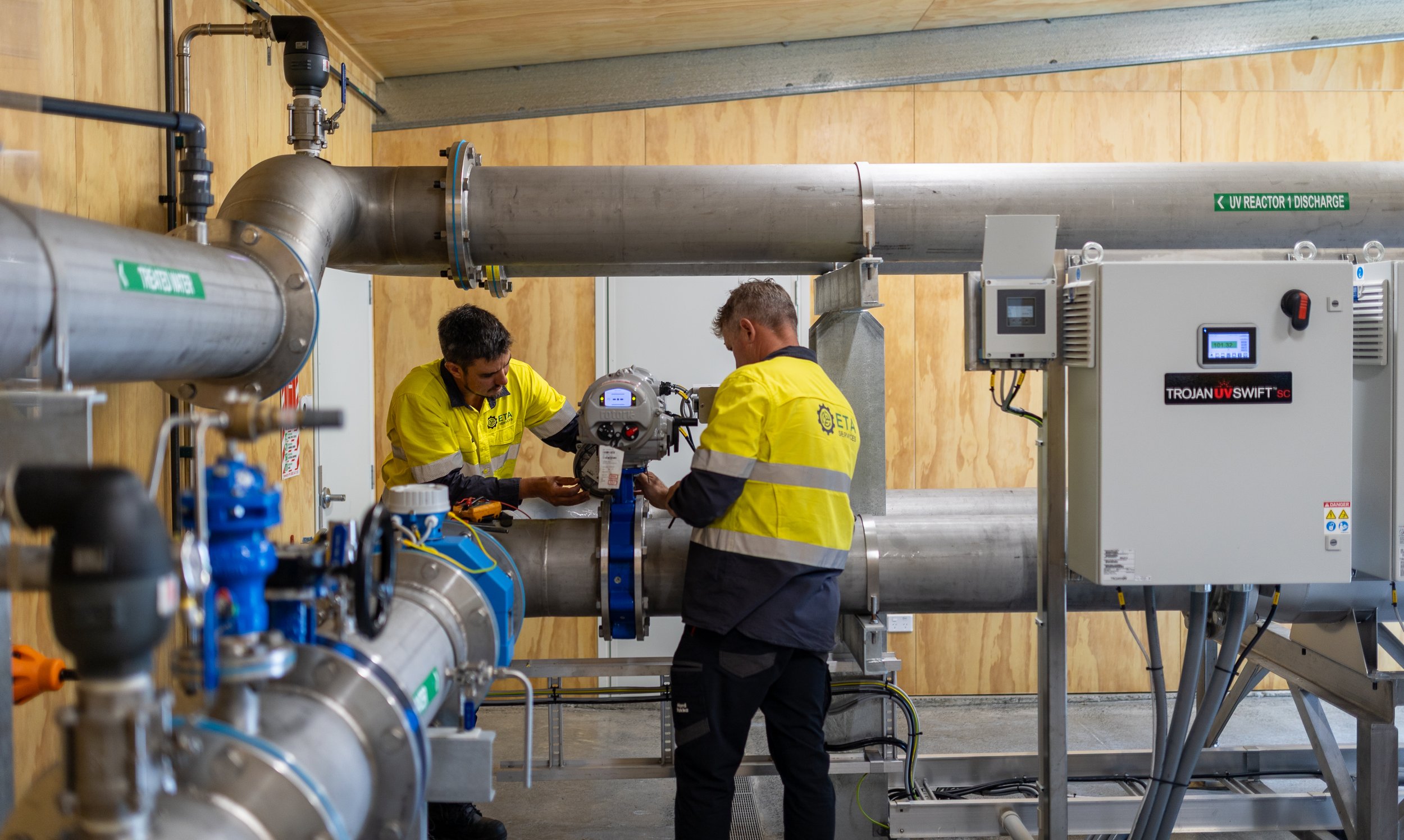 Two workers in yellow safety shirts working on industrial equipment with large pipes and control panels in a utility room.