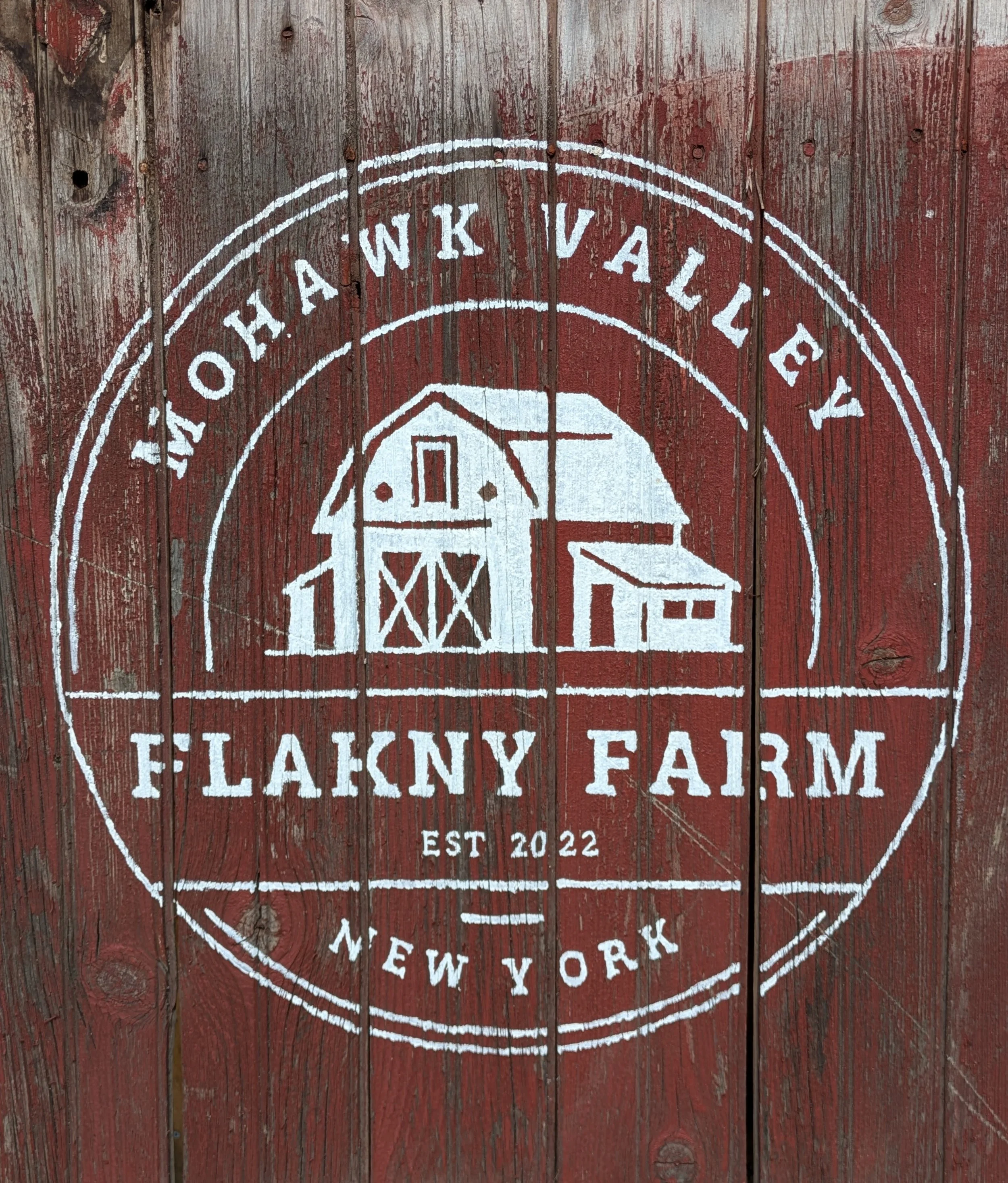 A round mural painted in white on a weathered red wooden fence, featuring a barn, silo, and farmhouse with the words 'FLAKNY FARM' along with 'EST 2022' at the bottom.
