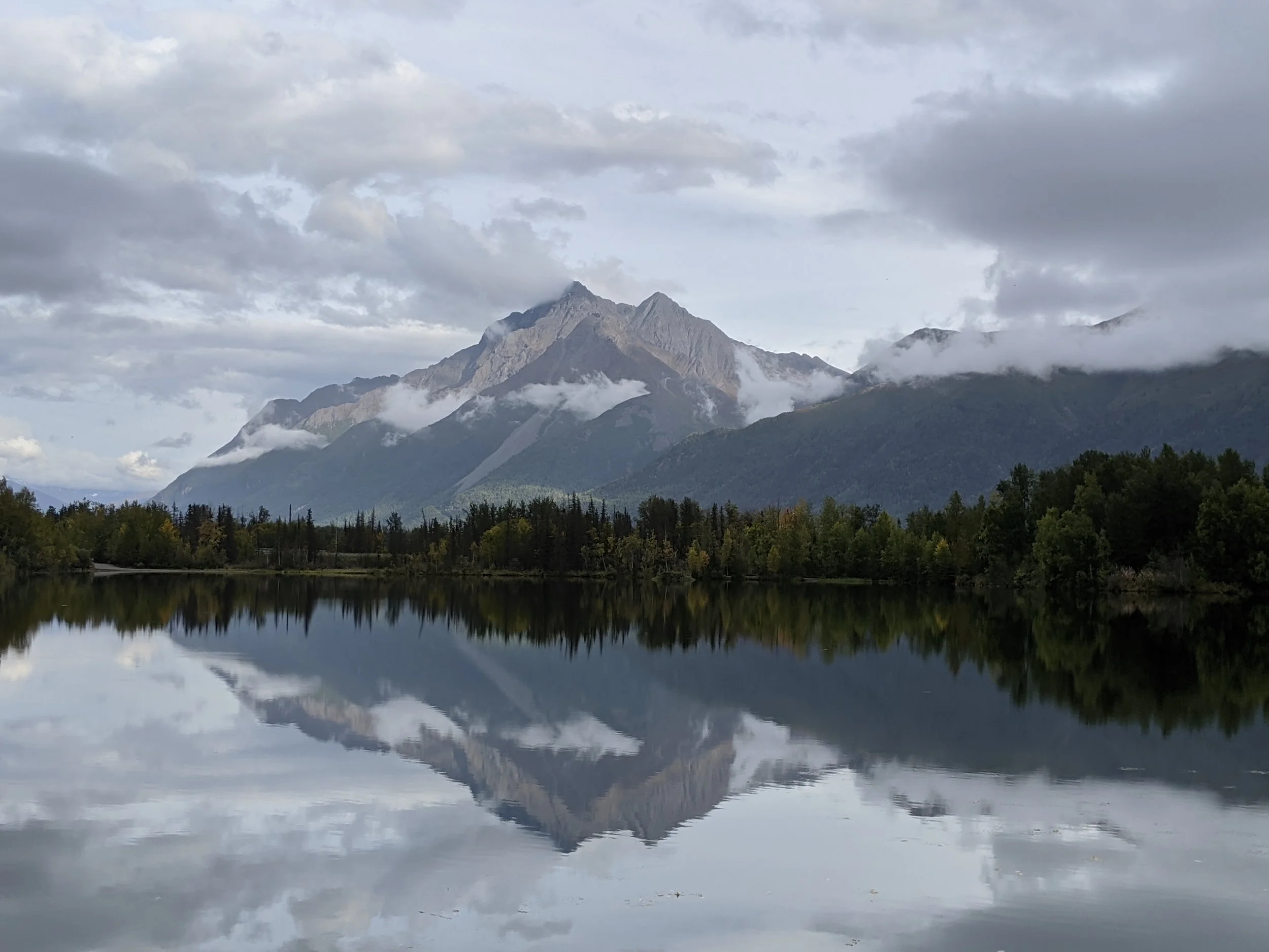 Mountain range with gray clouds overhead, reflected in a calm lake below surrounded by forest.