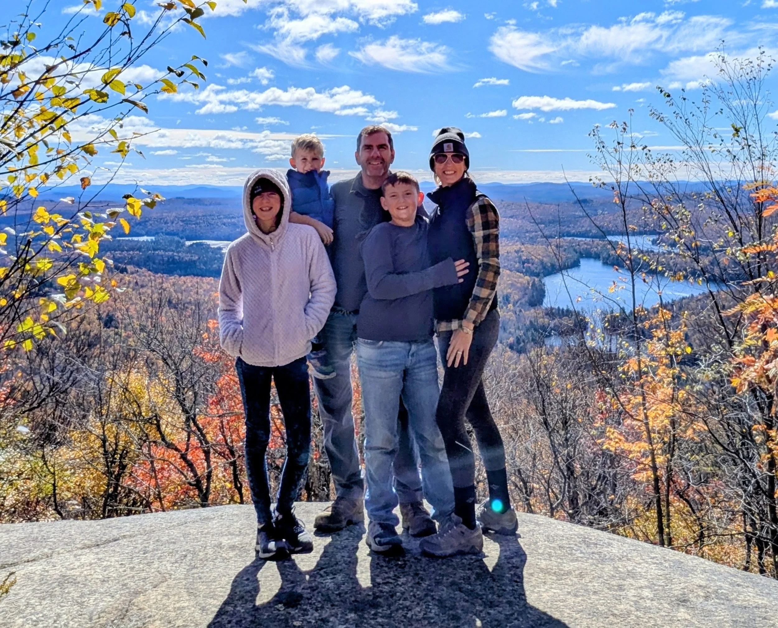 Our family of five, standing outdoors on a sunny day with a scenic view of mountains, a river, and a blue sky with clouds. They are dressed in casual fall clothing, smiling and close together, surrounded by autumn foliage.