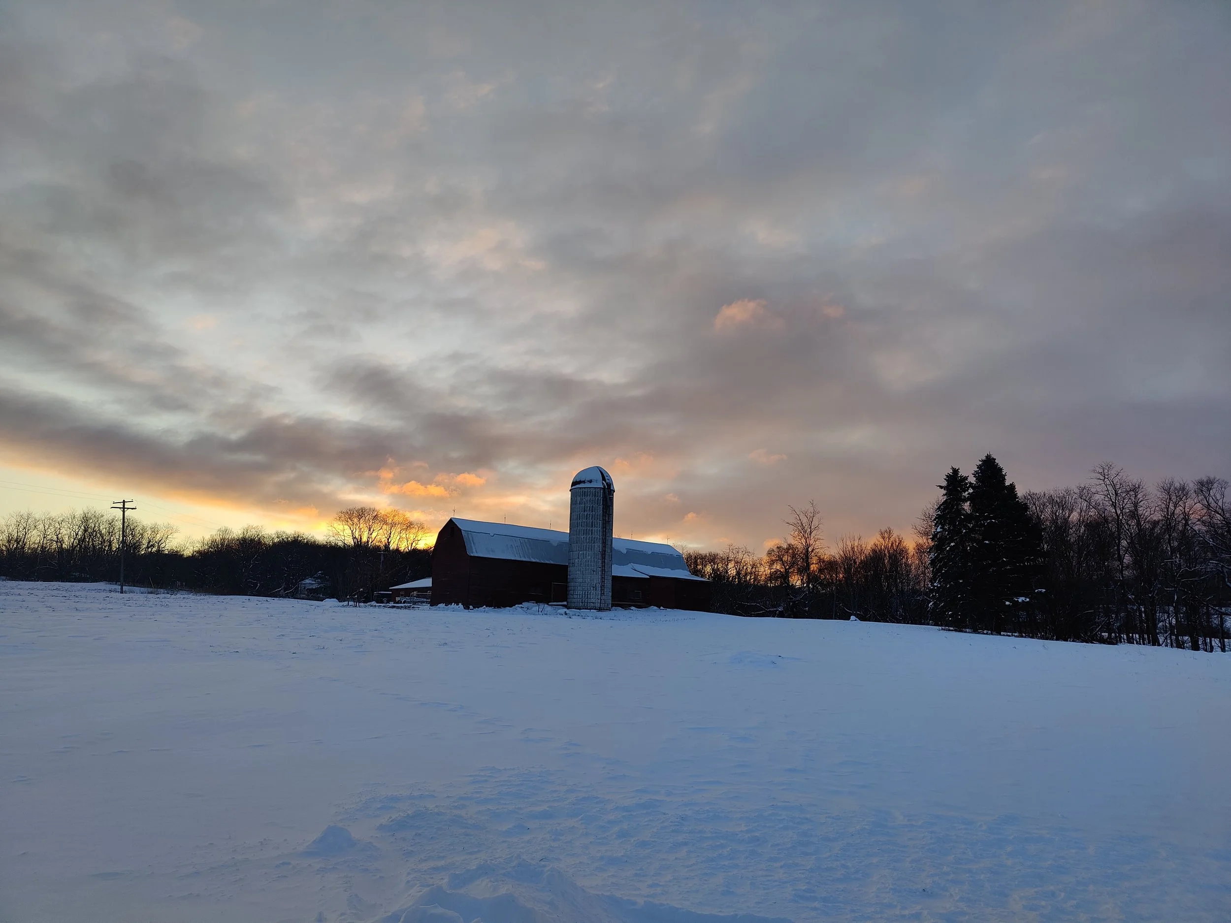 A rural farm scene at sunset with snow on the ground, a barn, a silo, and trees under a cloudy sky.