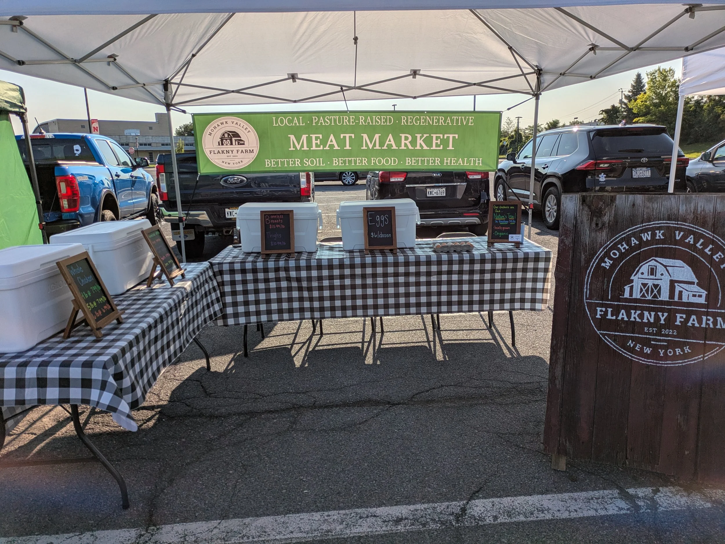 Outdoor market stall with a green sign reading 'Local, pasture-raised, regenerative meat market, better soil, better food, better health' in a parking lot, with cars parked behind and a building in the background.