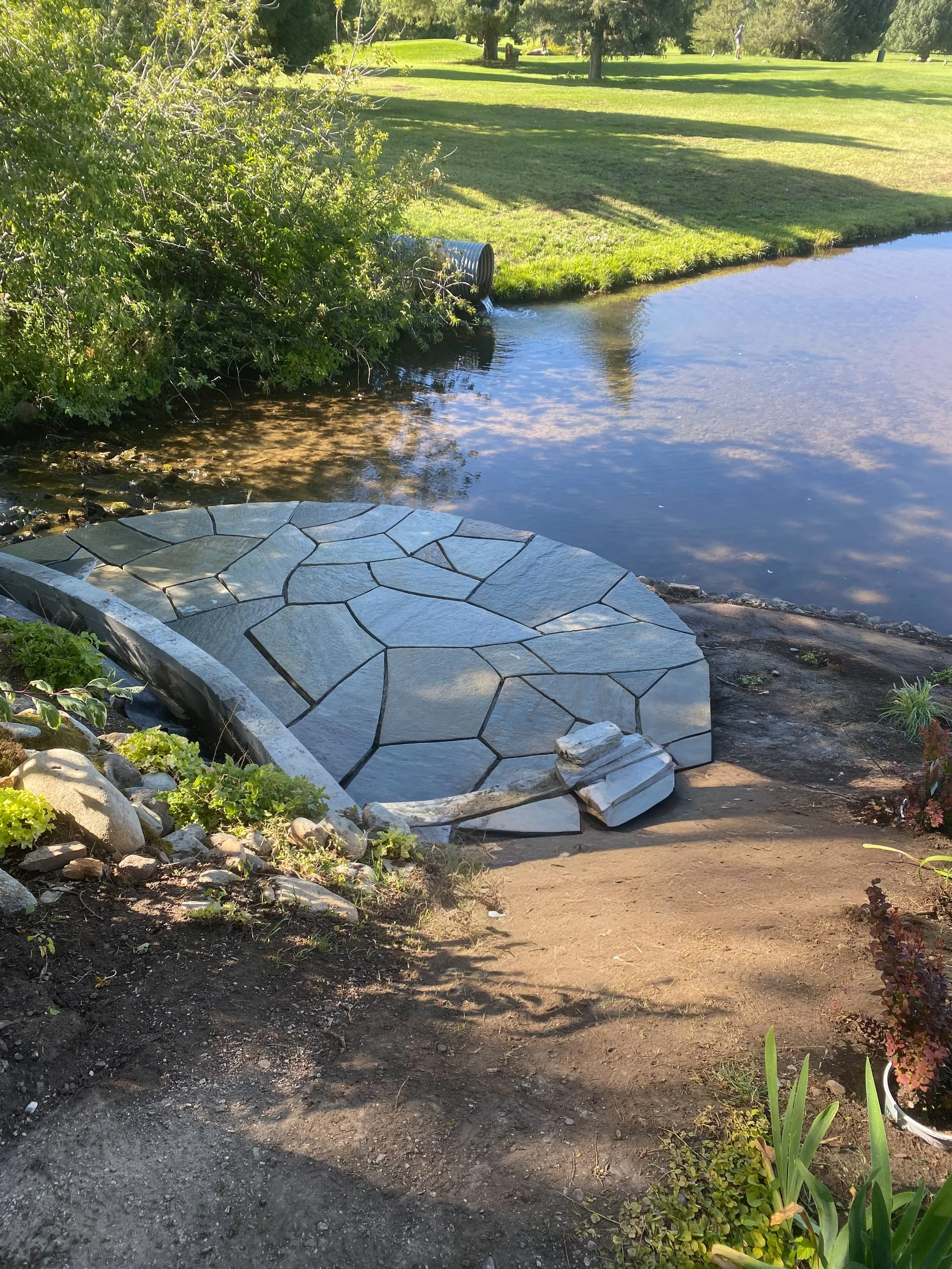 A small stone patio located at the edge of a pond, surrounded by dirt and plants, with a grassy area and trees in the background.