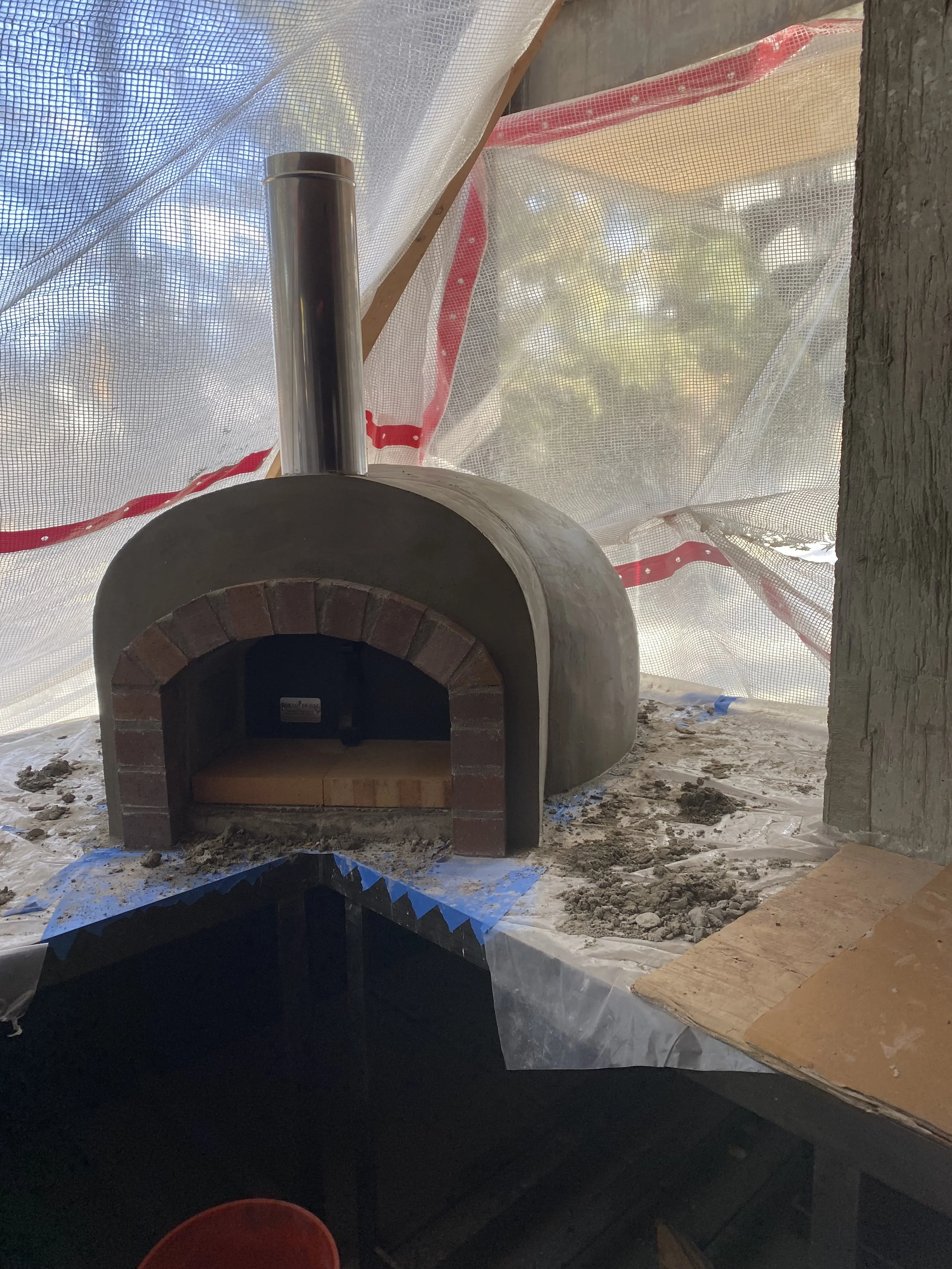 Small outdoor brick pizza oven with a silver chimney, surrounded by construction materials and a plastic tarp covering the area.