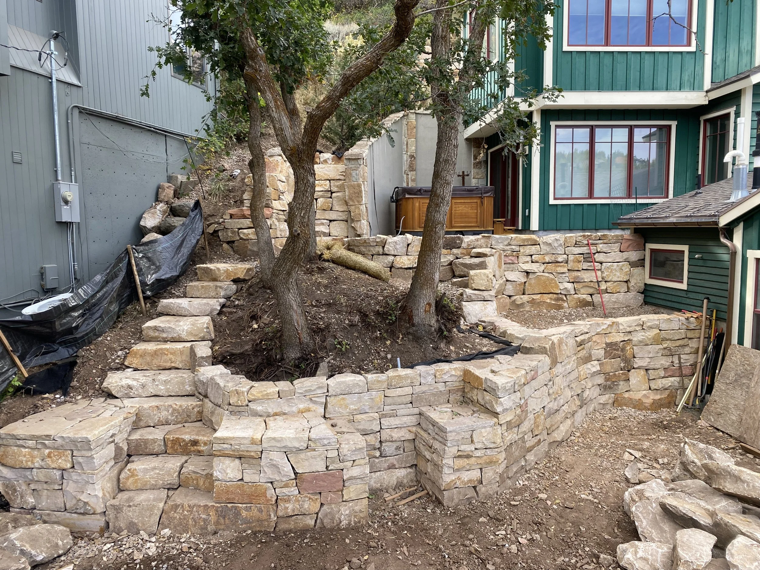 Construction site with partially built stone retaining wall around a yard with two trees, a hot tub, and a green house with large windows.