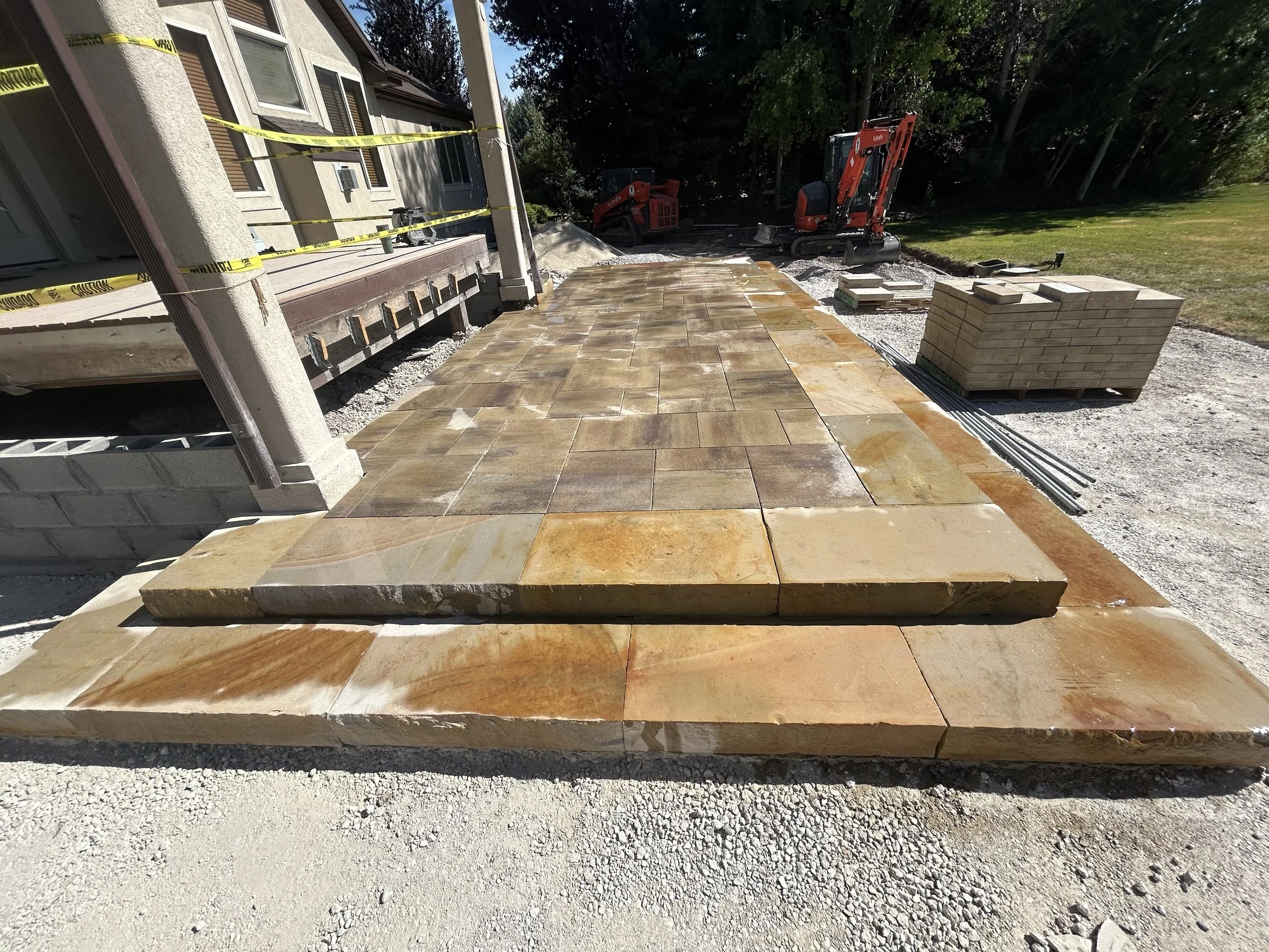 Outdoor construction site with a partially completed stone patio, stacks of stones, construction equipment, and caution tape.