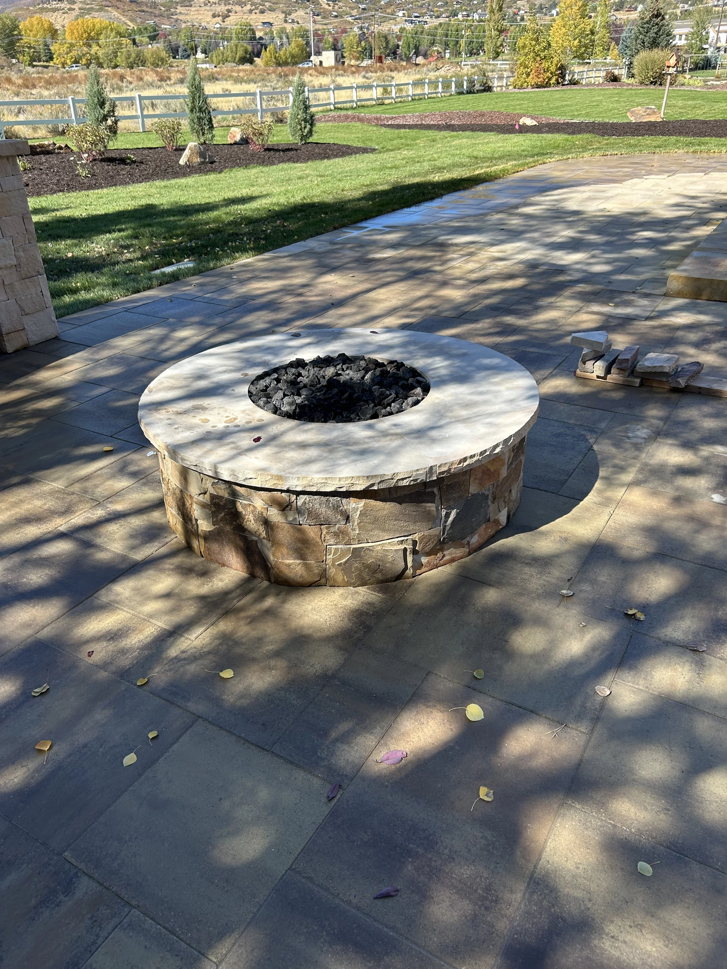 Stone fire pit on a patio with a circular top filled with black stones, surrounded by pavers, with a yard and trees in the background.