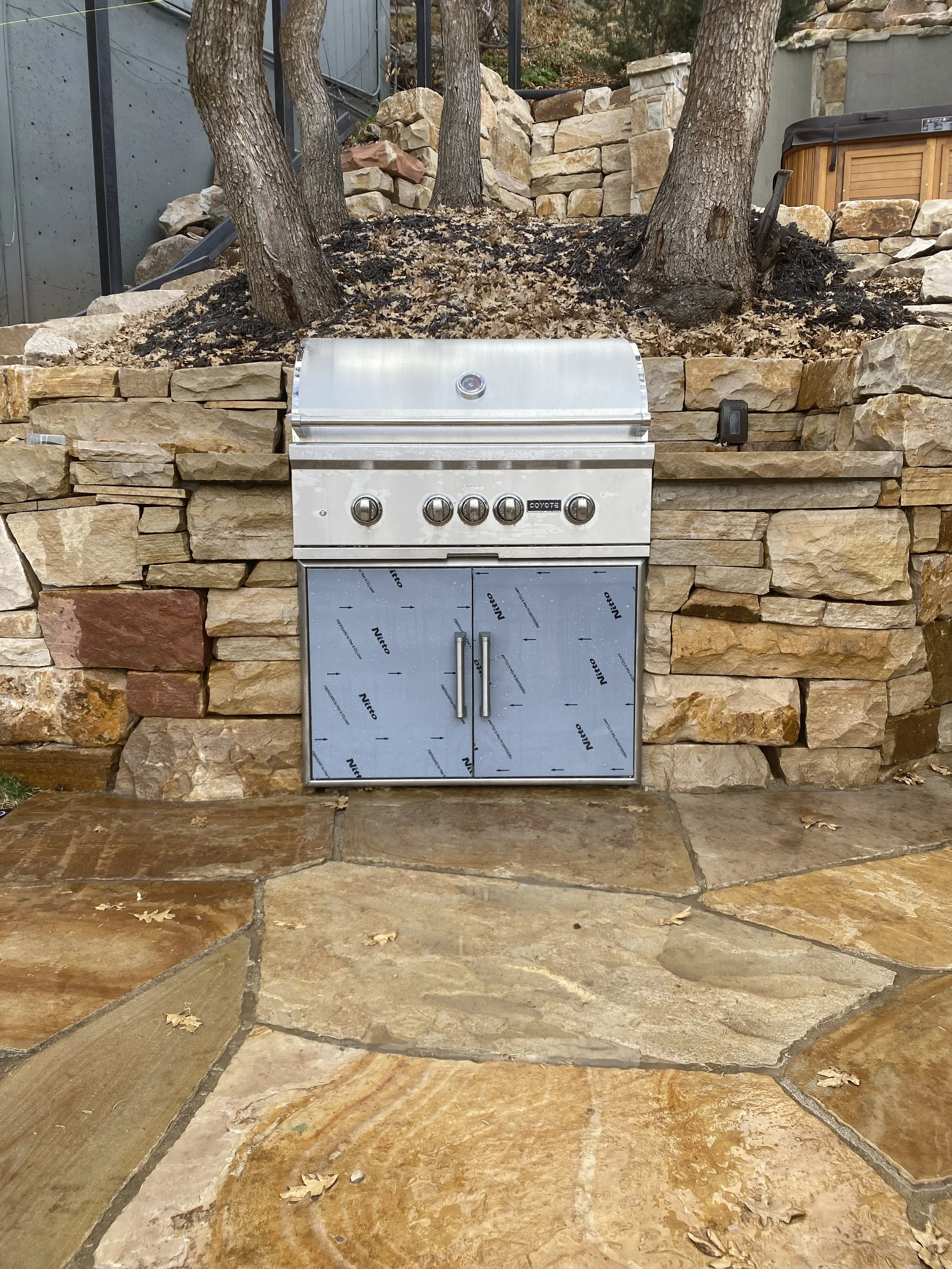 Outdoor built-in stainless steel grill with cabinet, set against a stone wall with trees and dry leaves.