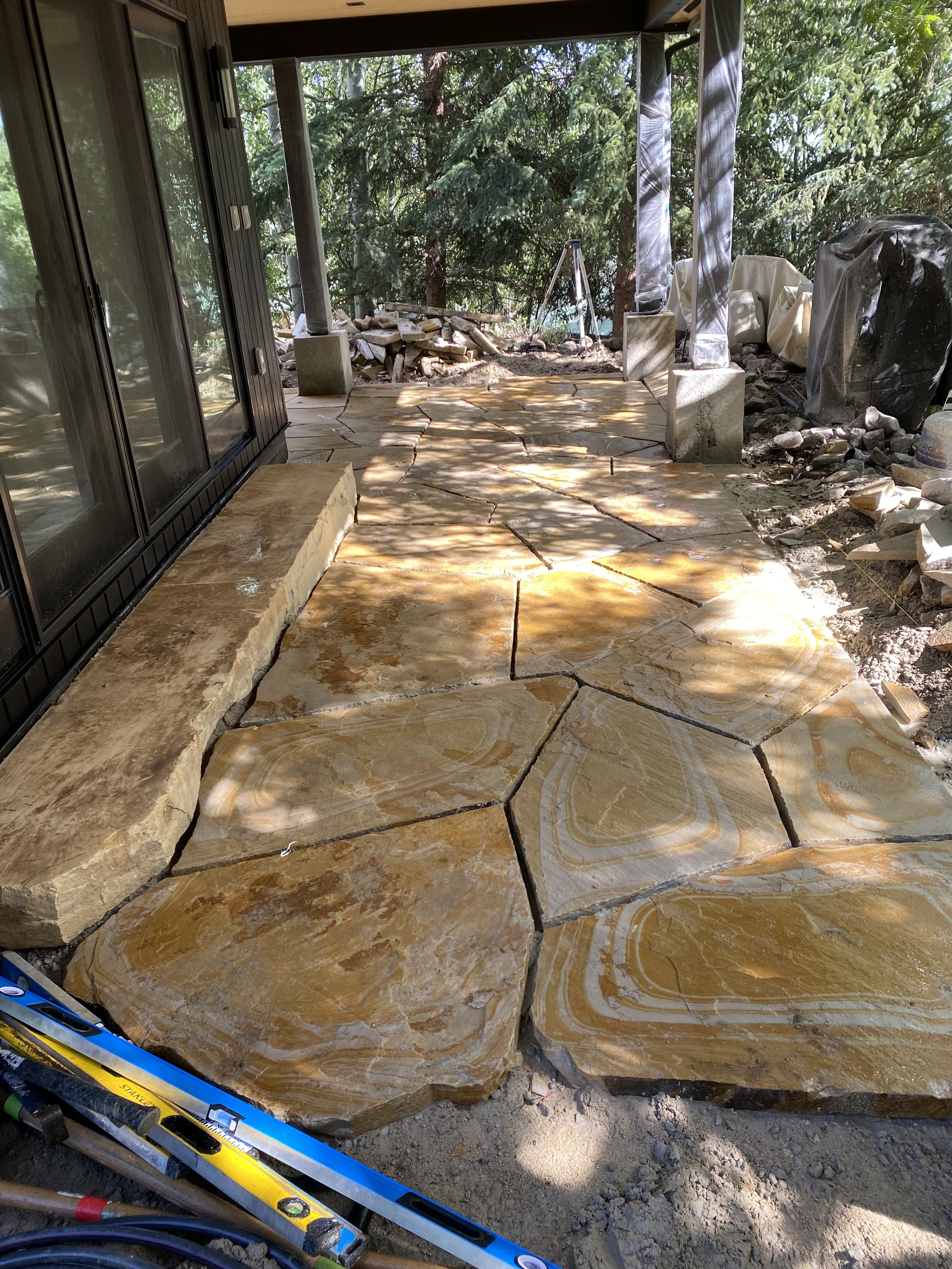 Under construction outdoor patio with large irregular yellowish sandstone slabs being installed on the ground, surrounded by tools and construction materials.