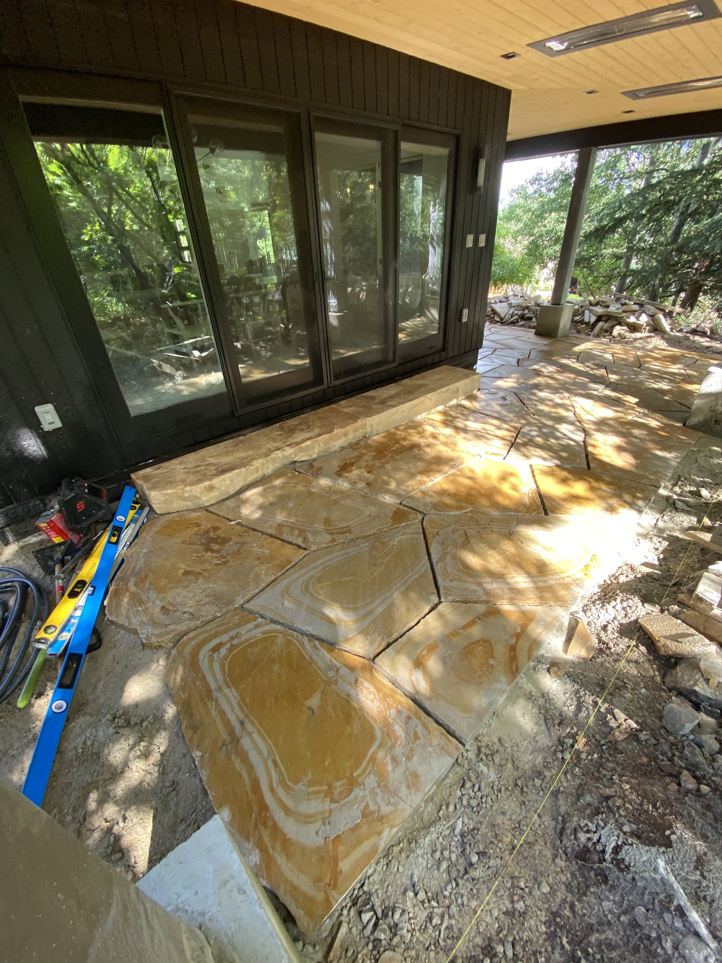 Outdoor patio area with large, irregularly shaped sandstone slab tiles being installed, next to a glass sliding door and surrounded by trees.
