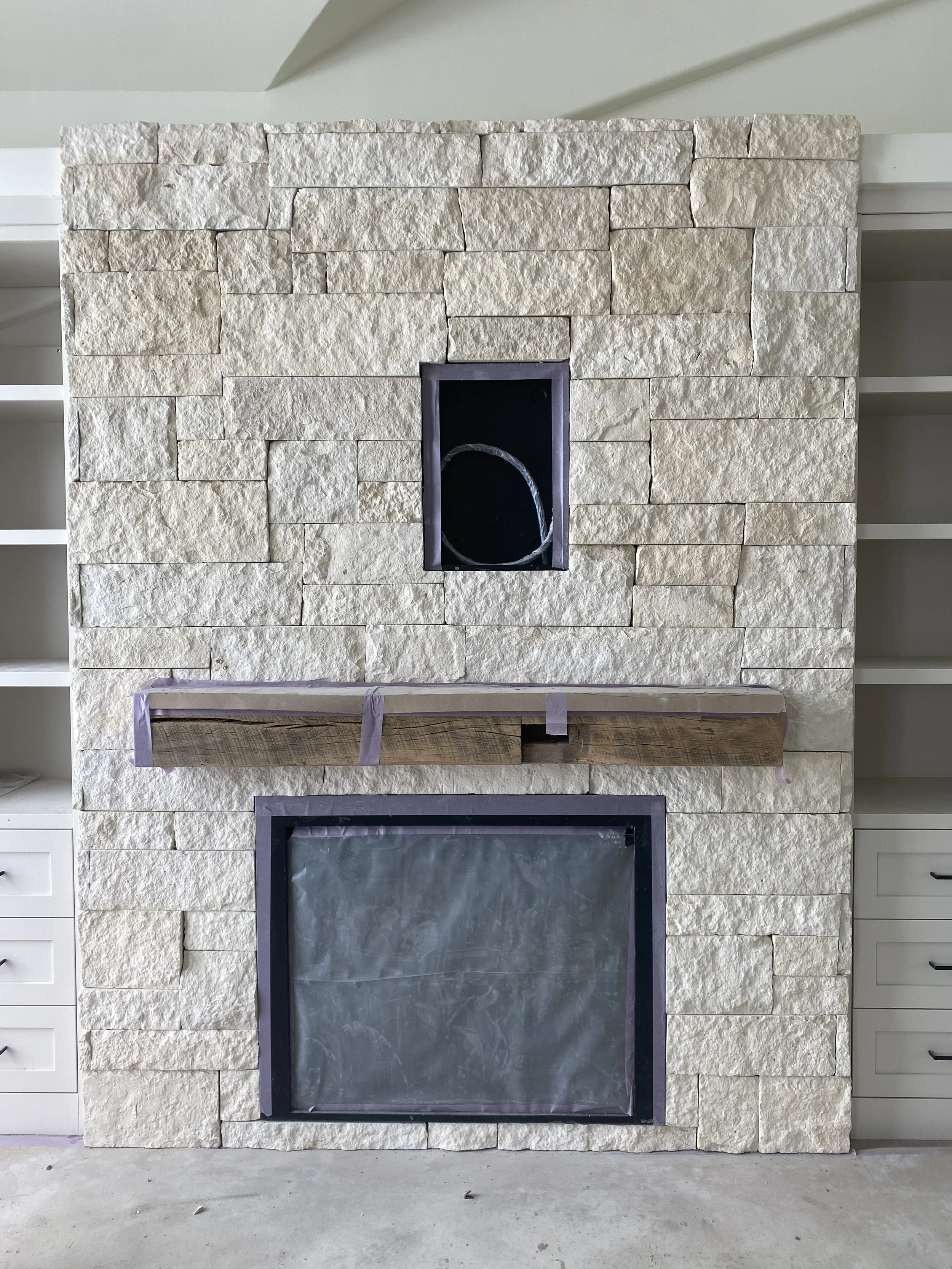 A stone fireplace under construction with a rectangular opening at the bottom covered with plastic, a wooden mantel with purple tape, and a smaller rectangular opening at the top with black trim, all flanked by built-in white cabinets.