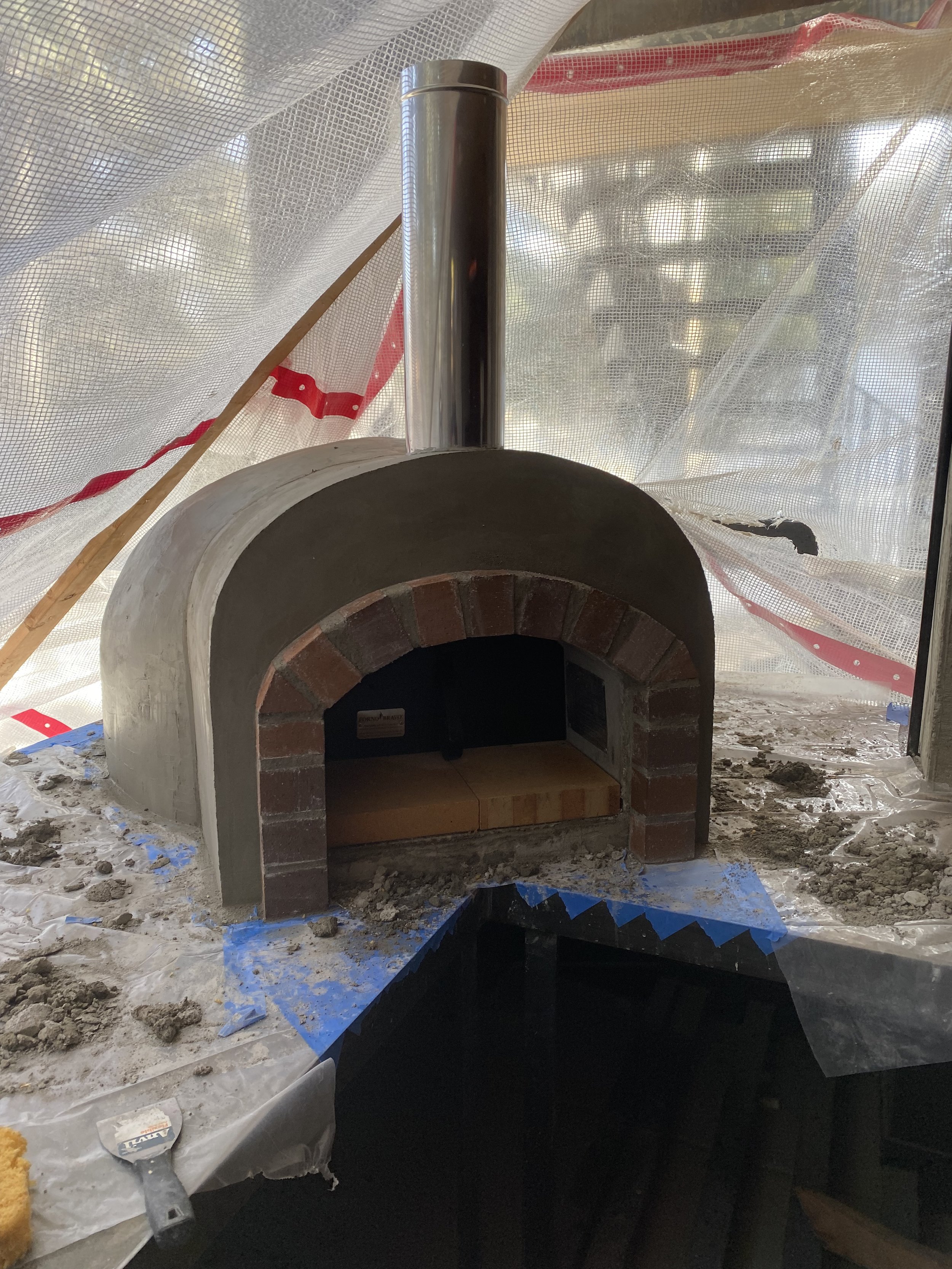 Small brick and concrete pizza oven under construction, with a metal chimney pipe on top, inside a covered area with plastic sheeting and construction materials around.