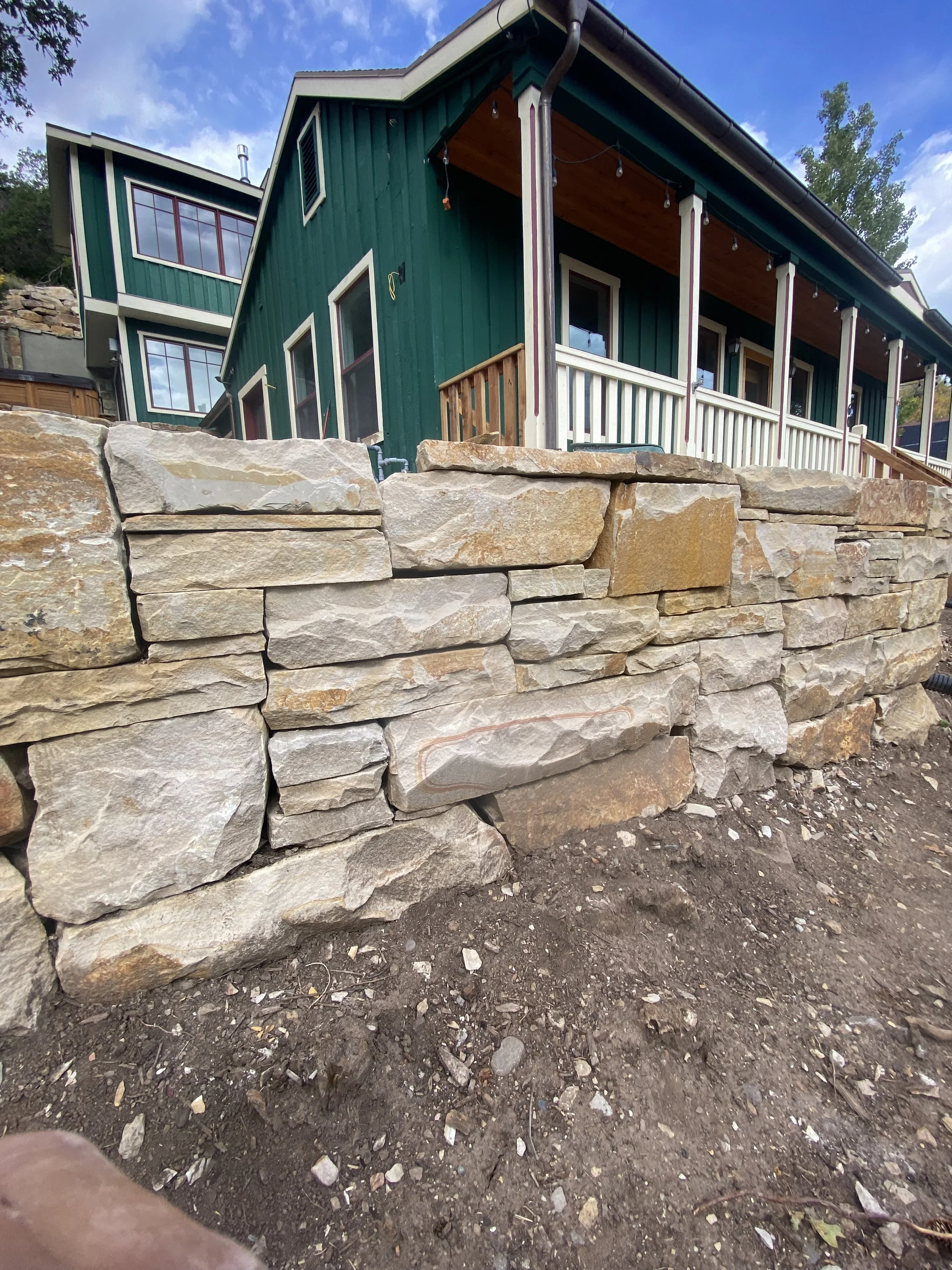 A stone retaining wall in front of a green house with white trim and a porch with string lights.