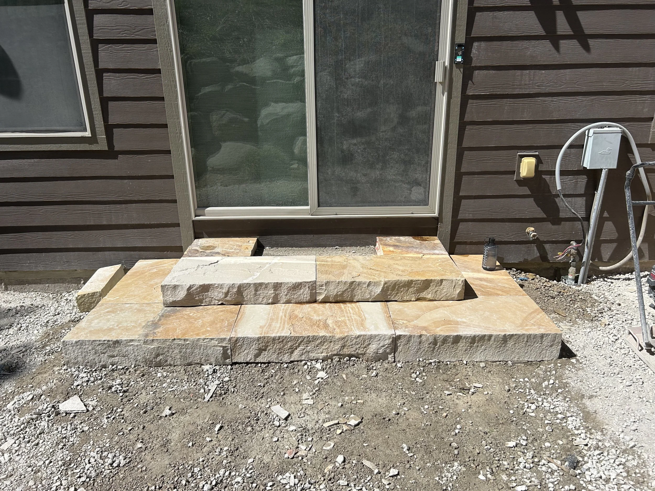 Stone steps under construction in front of a glass sliding door, with brown exterior siding and construction materials nearby.