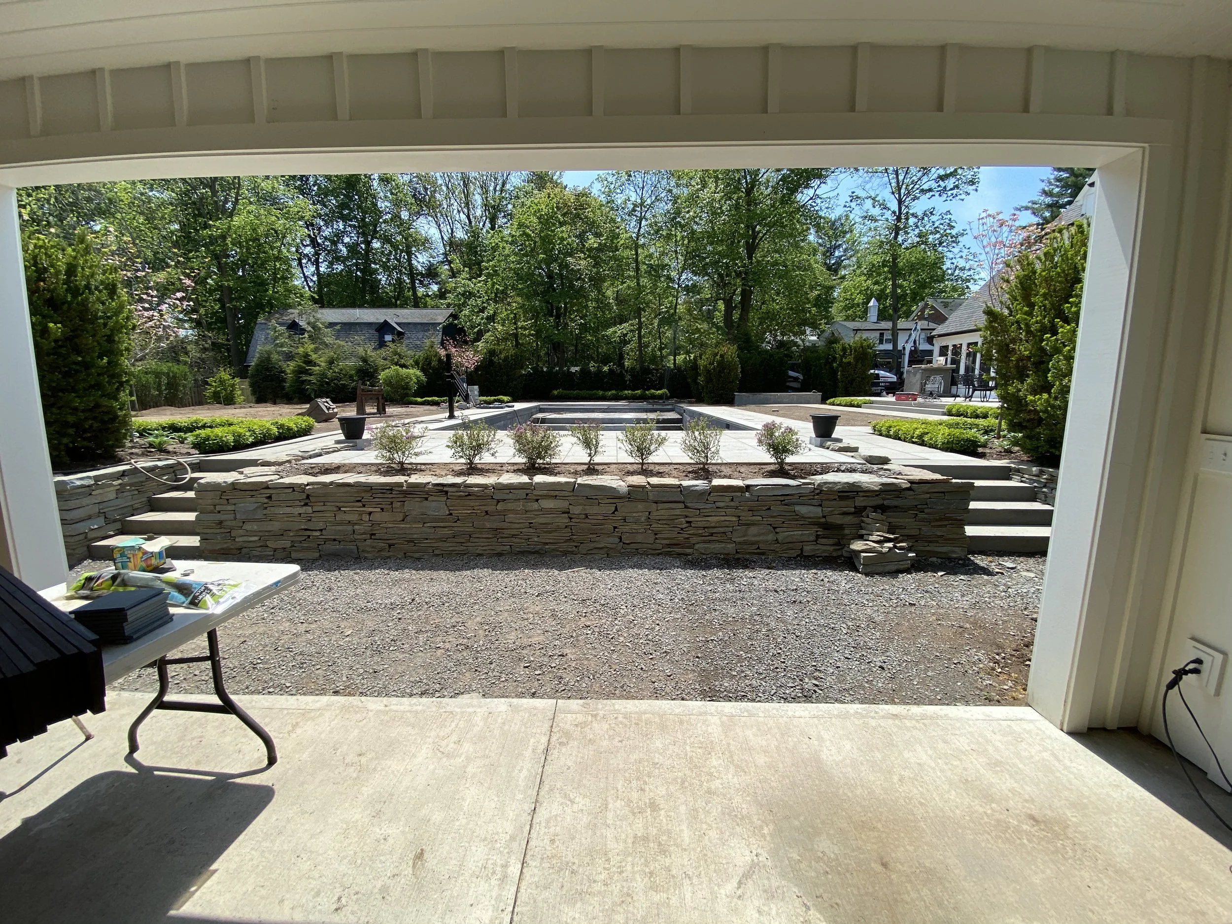 View from a covered patio looking onto a landscaped backyard with stone steps and retaining wall, small bushes, trees, and neighboring houses in the background.