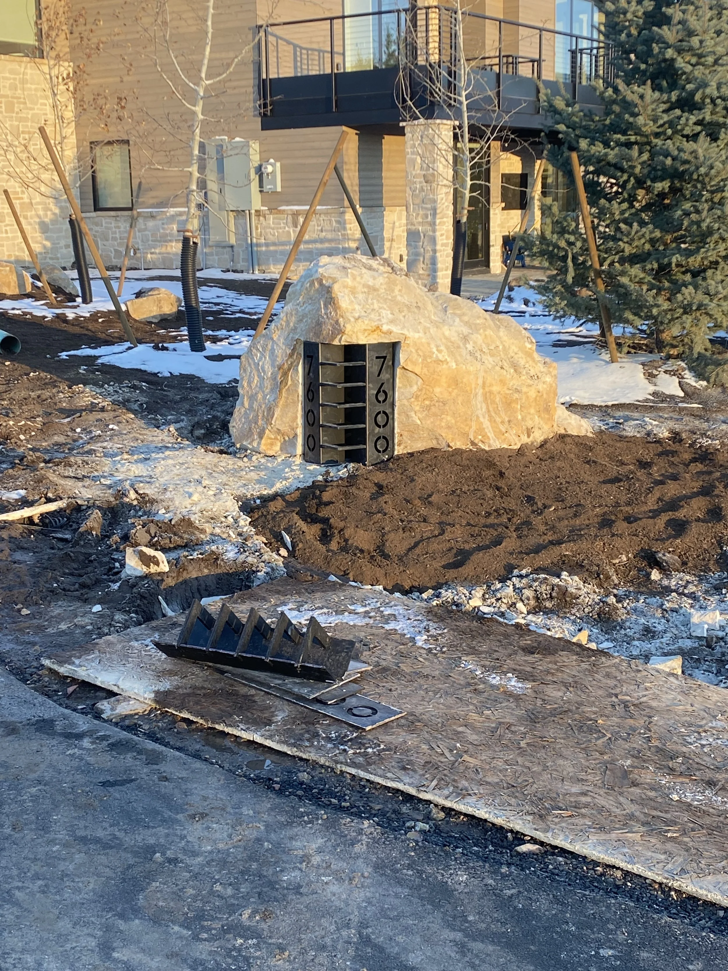 A large rock with a thermometer sign on it showing temperatures from 60 to 70 degrees Fahrenheit, surrounded by construction materials and dirt, with patches of snow and a building with a balcony in the background.