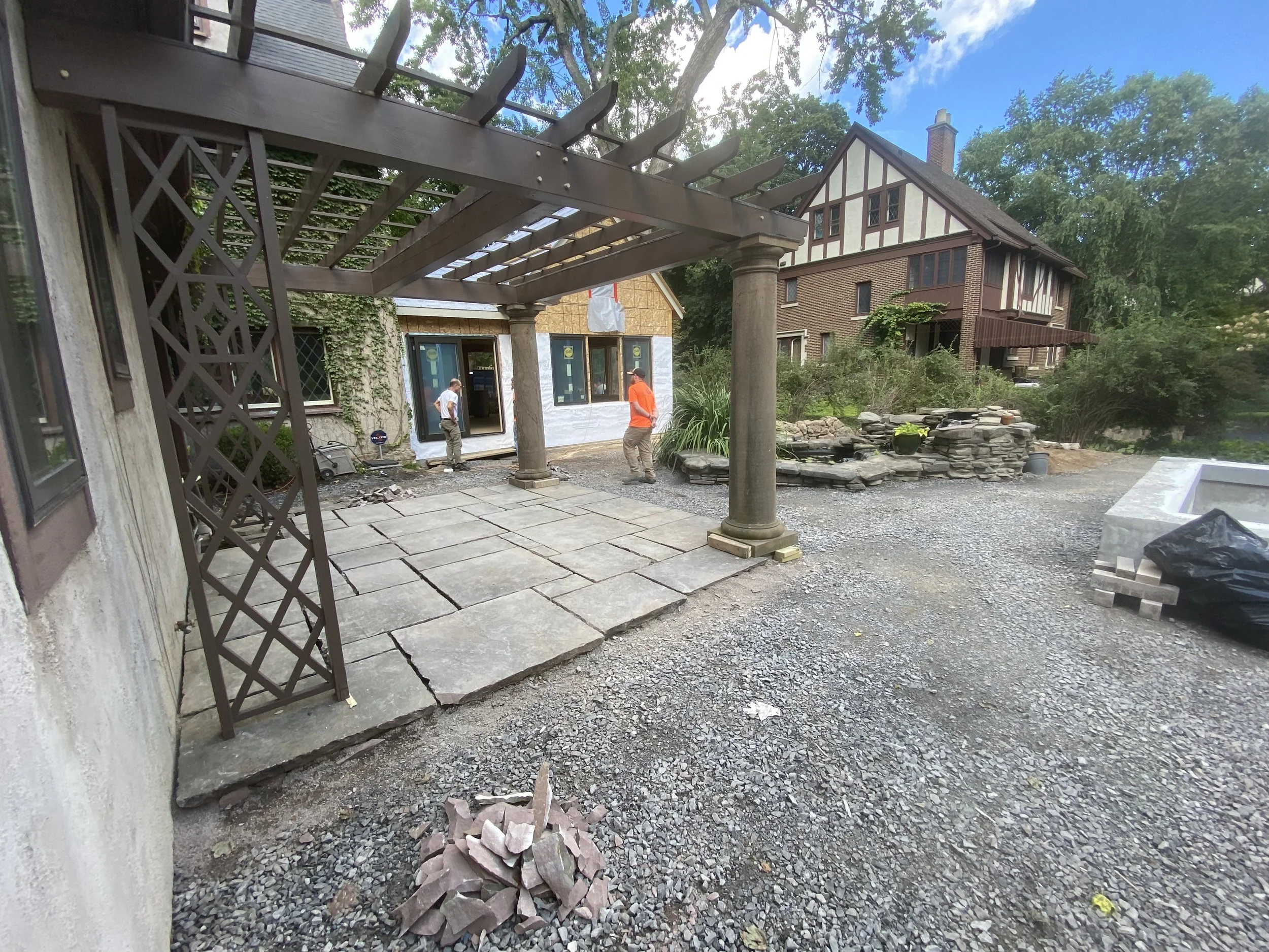 Construction site for a patio with large stone tiles being installed in backyard. Two workers are present, one in an orange shirt and another in a gray shirt. There is a pergola with columns and a lattice panel, a landscaped rock feature, and a house