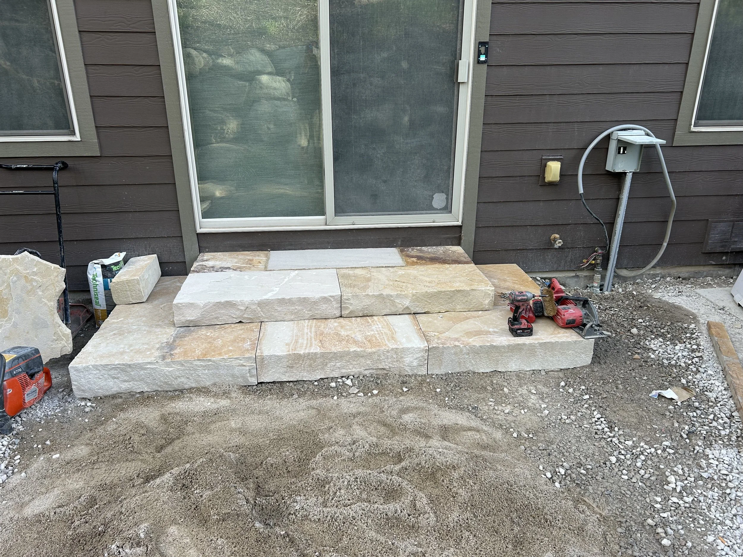 Construction site showing a partially built stone front step in front of a sliding glass door on a house with brown siding. Construction tools and materials, including a cordless drill, in the foreground.