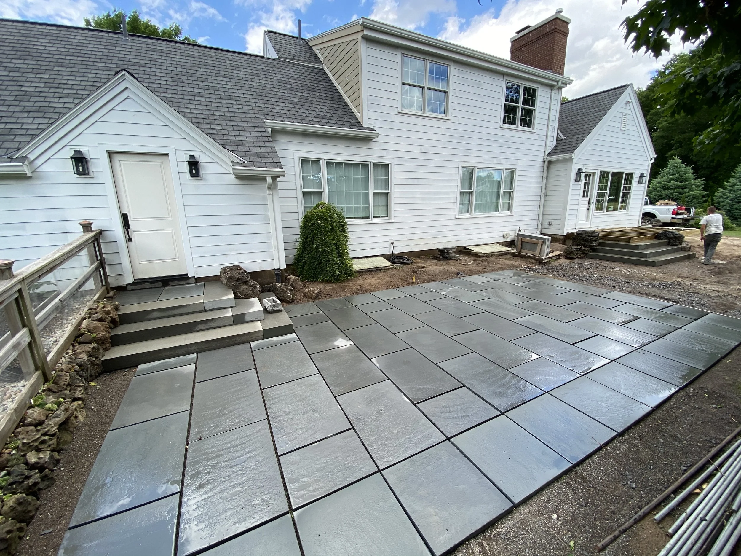 Newly paved stone patio area in the backyard of a white house with steps leading up to the house entrance. There are construction materials and workers present, with a truck parked in the background.
