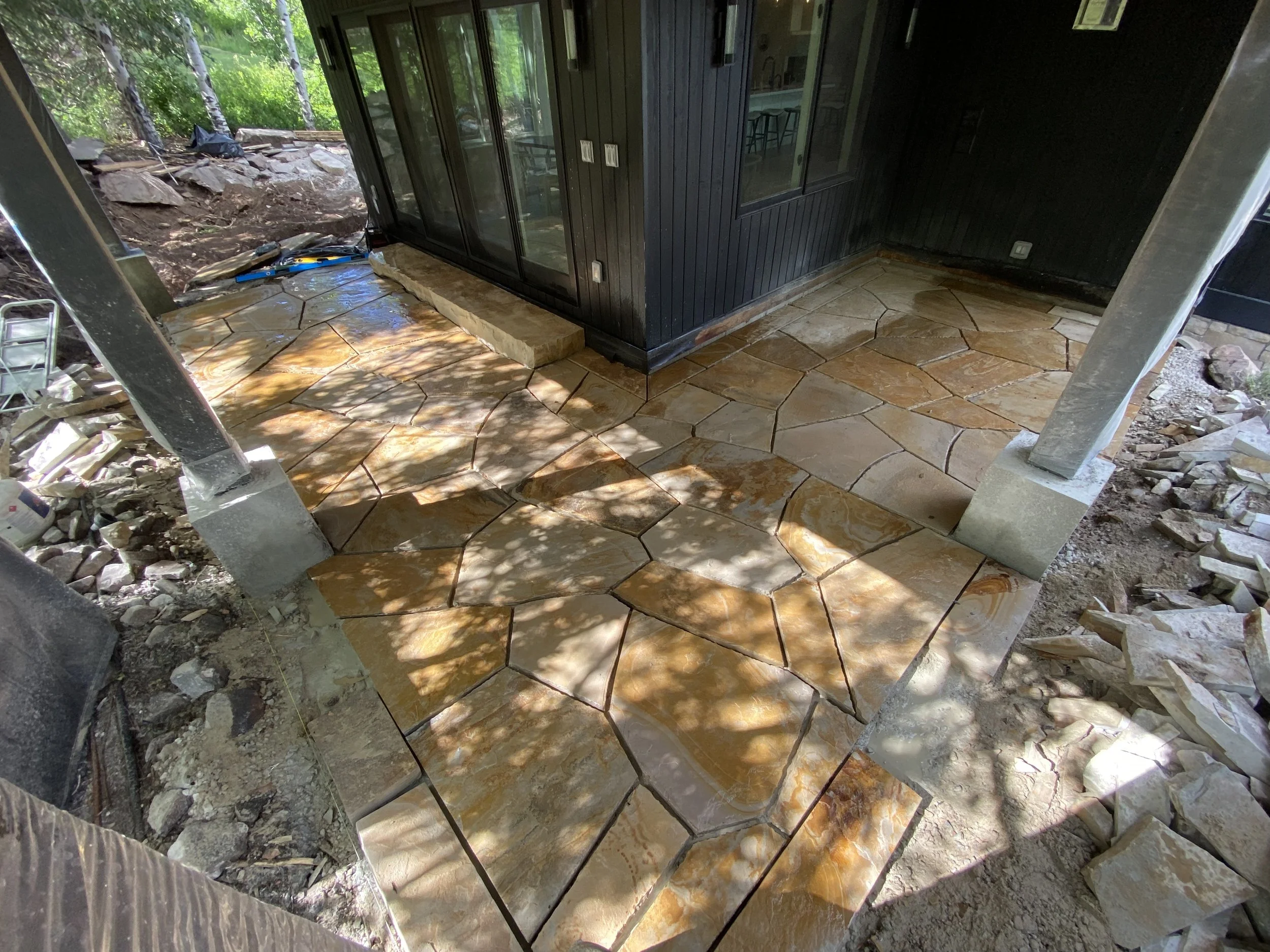 Stone patio being installed outside a house with black siding, with sunlight and shadows from trees, and construction tools nearby.