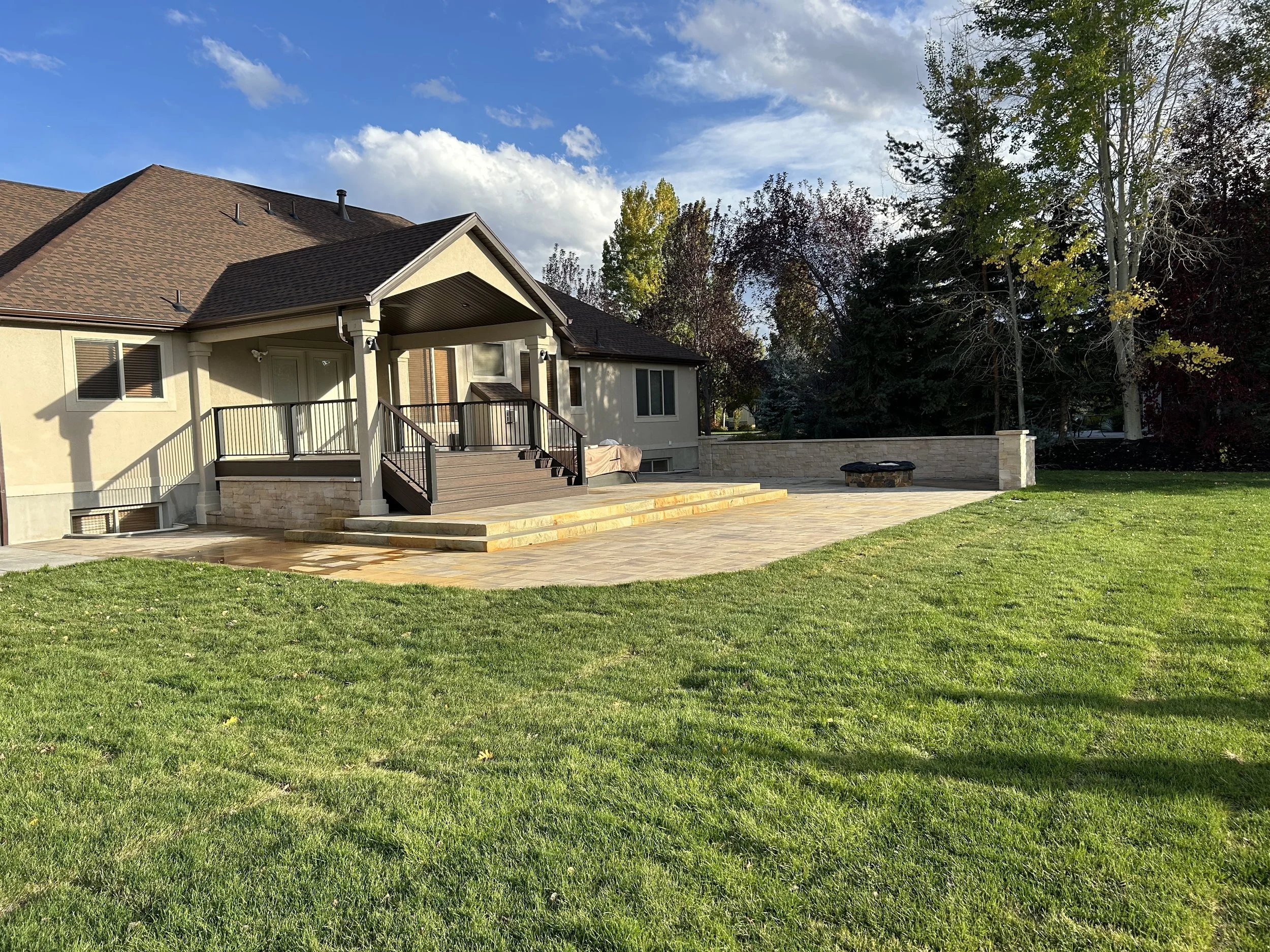 Backyard patio area with a wooden deck, stone steps, and a fire pit, surrounded by green grass and tall trees.