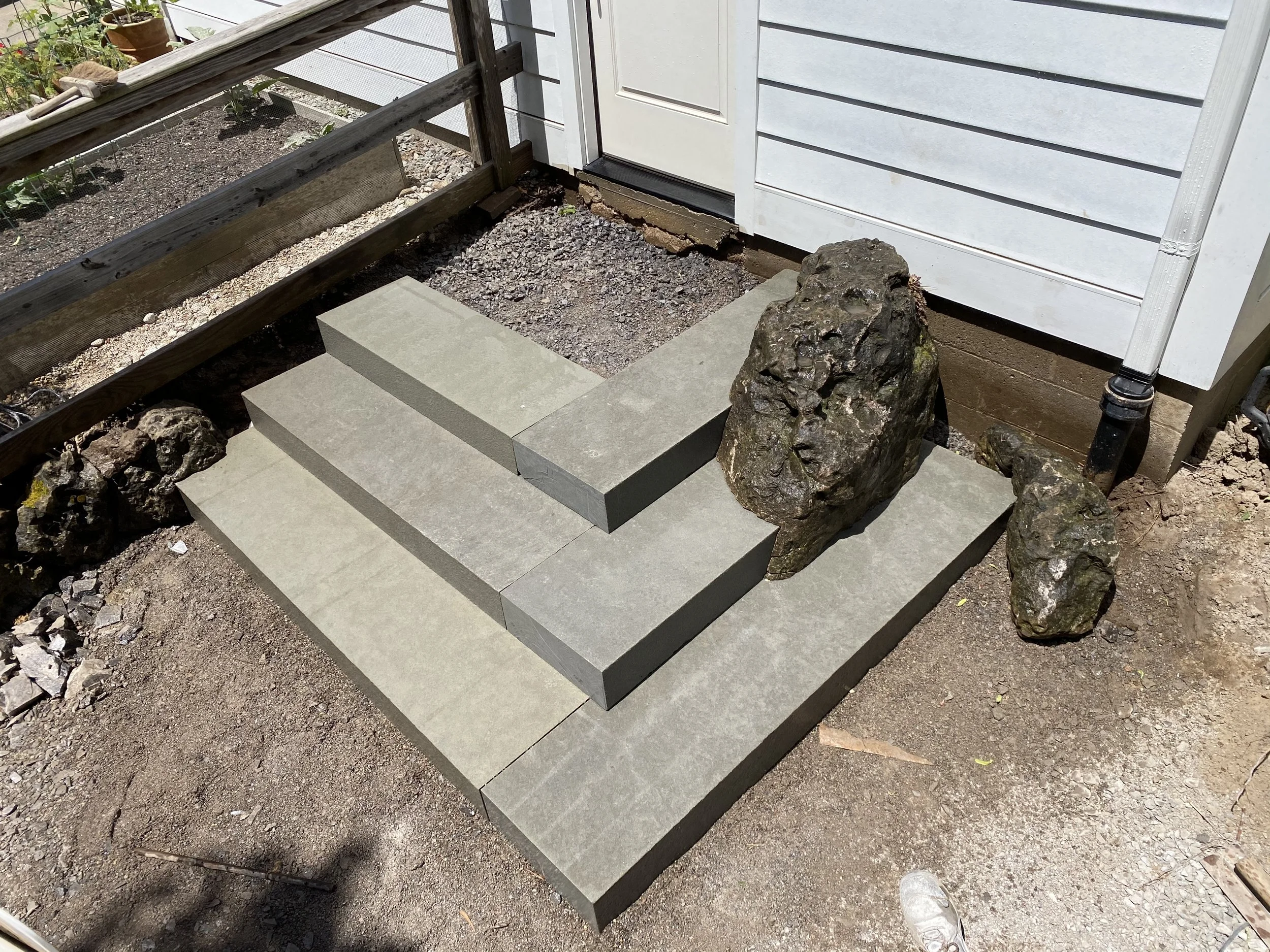 Stone stairs with three steps, placed next to large rocks, leading to a white door of a house with white siding, on a dirt surface.