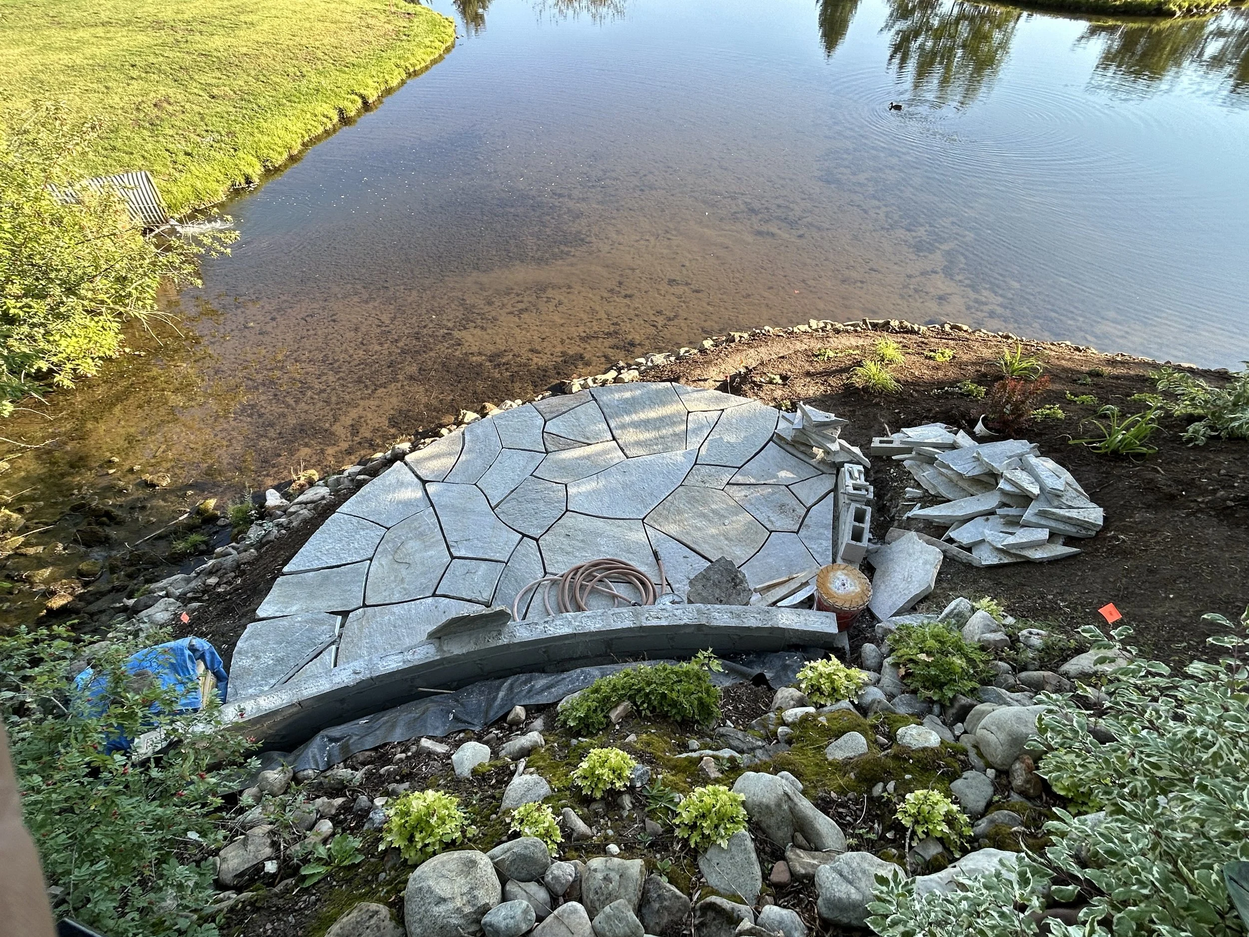 Under construction stepping stone pathway by a pond with water and green vegetation.