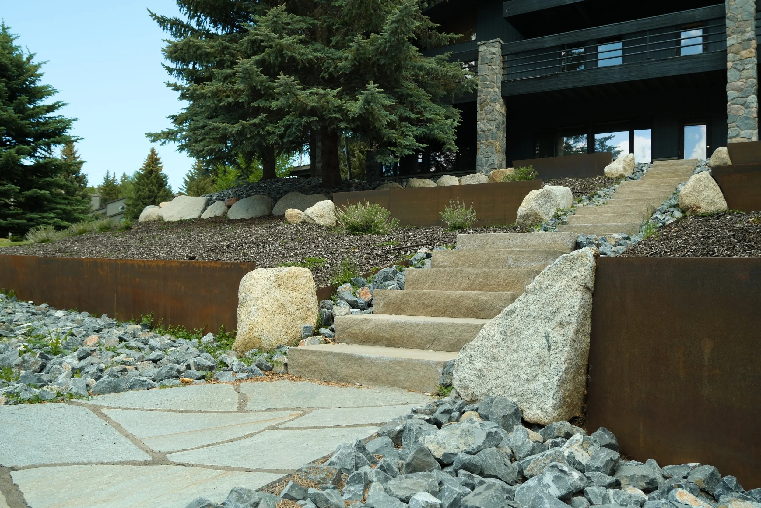 Newly constructed multi-story house with wooden siding, stone foundation, and balconies, adjacent to a dirt hillside with stone steps and a patio area.